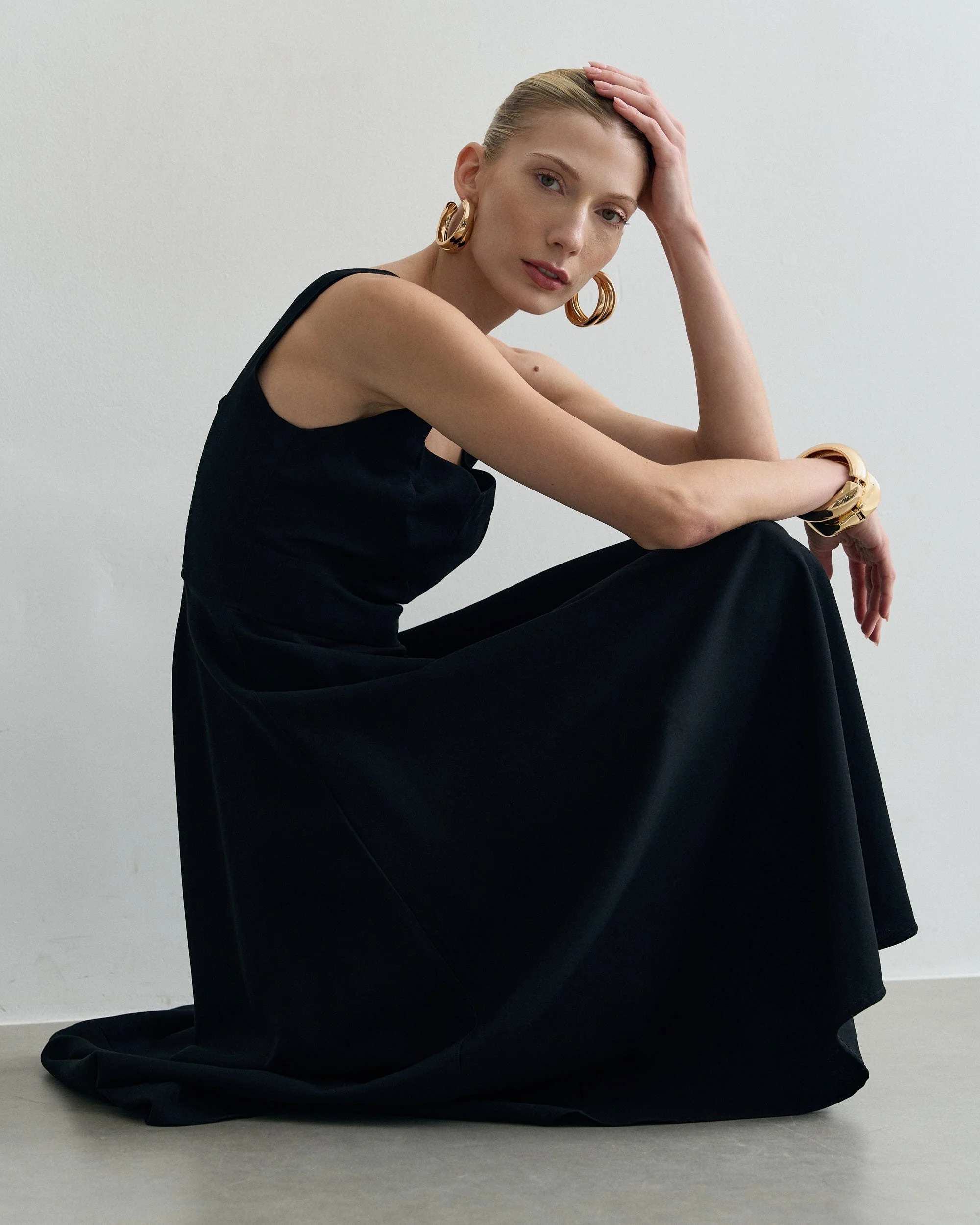 Mulher com pele clara, cabelo loiro preso, usando roupa preta, com grandes brincos dourados e bracelete dourado, sentada com os joelhos dobrados na frente de um fundo branco. Ela olha para a câmera com expressão serena.
