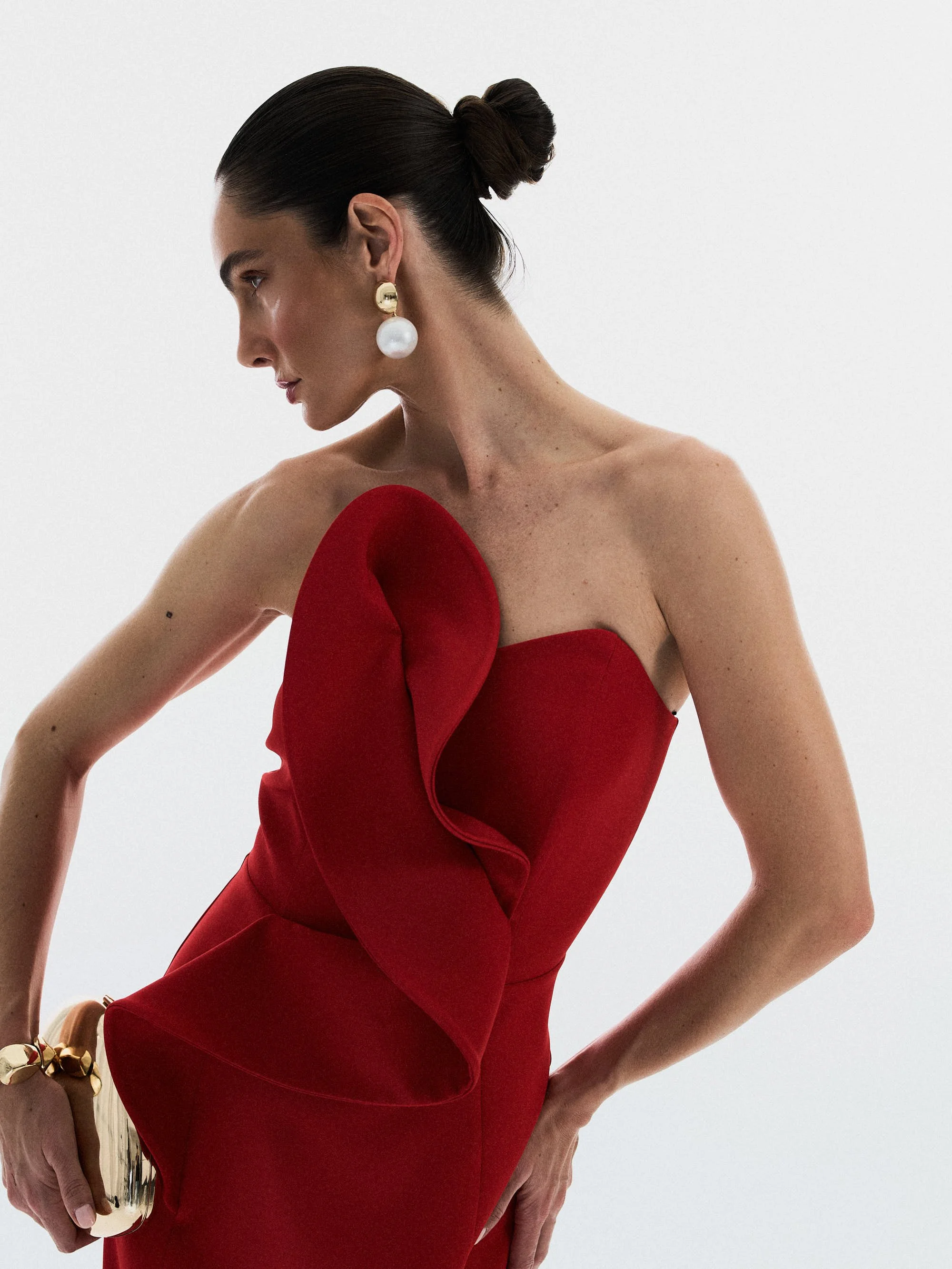 Mulher com cabelo preso em coque, usando brincos grandes de pérola e dourado, vestido vermelho com detalhe de babado na frente, segurando uma clutch dourada, contra fundo branco.