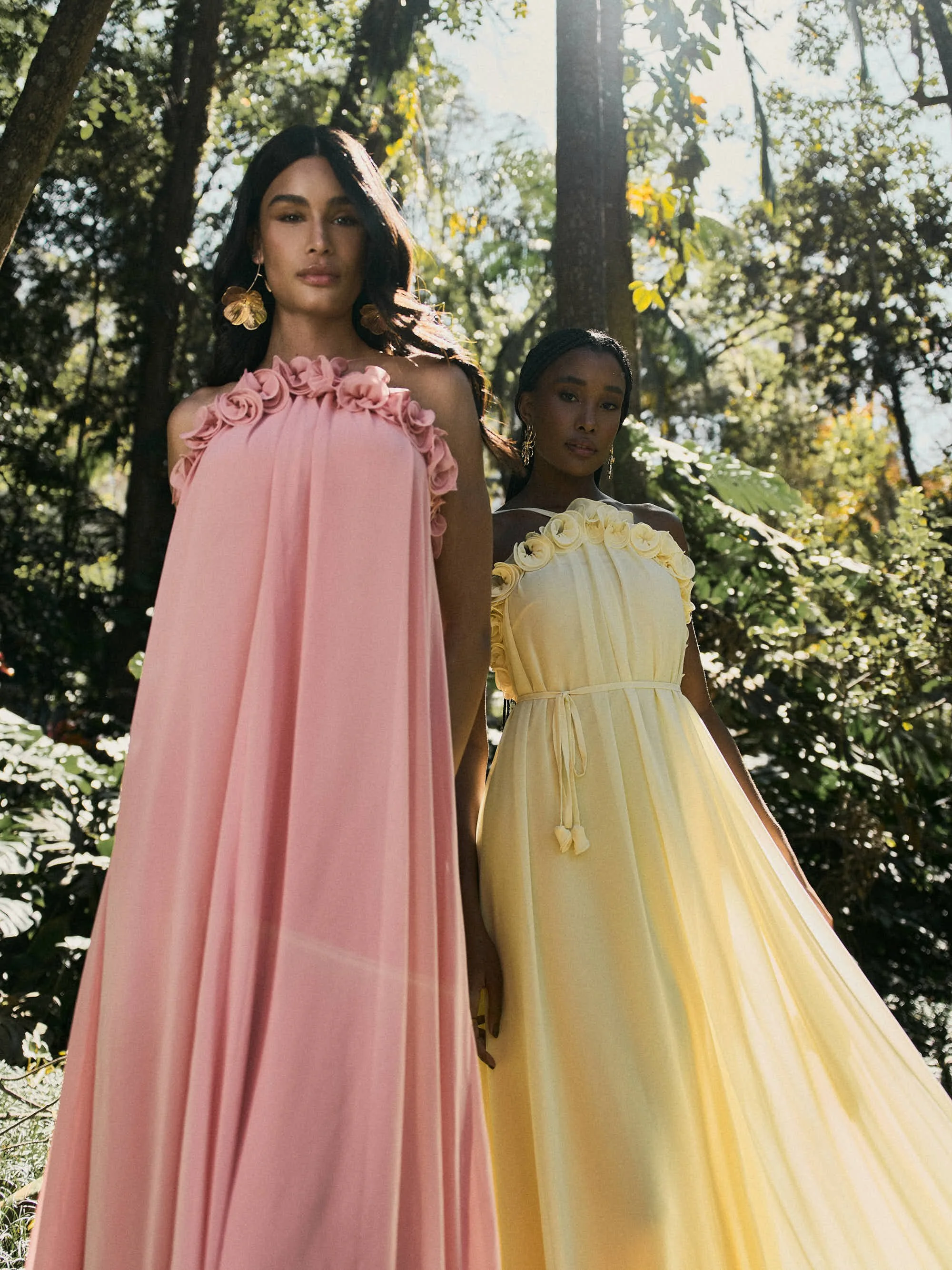 Duas mulheres em vestidos de gala pastel, uma rosa e outra amarela, com detalhes de rosas e pregas, posando em uma floresta com árvores altas e luz solar filtrando pelas folhas.