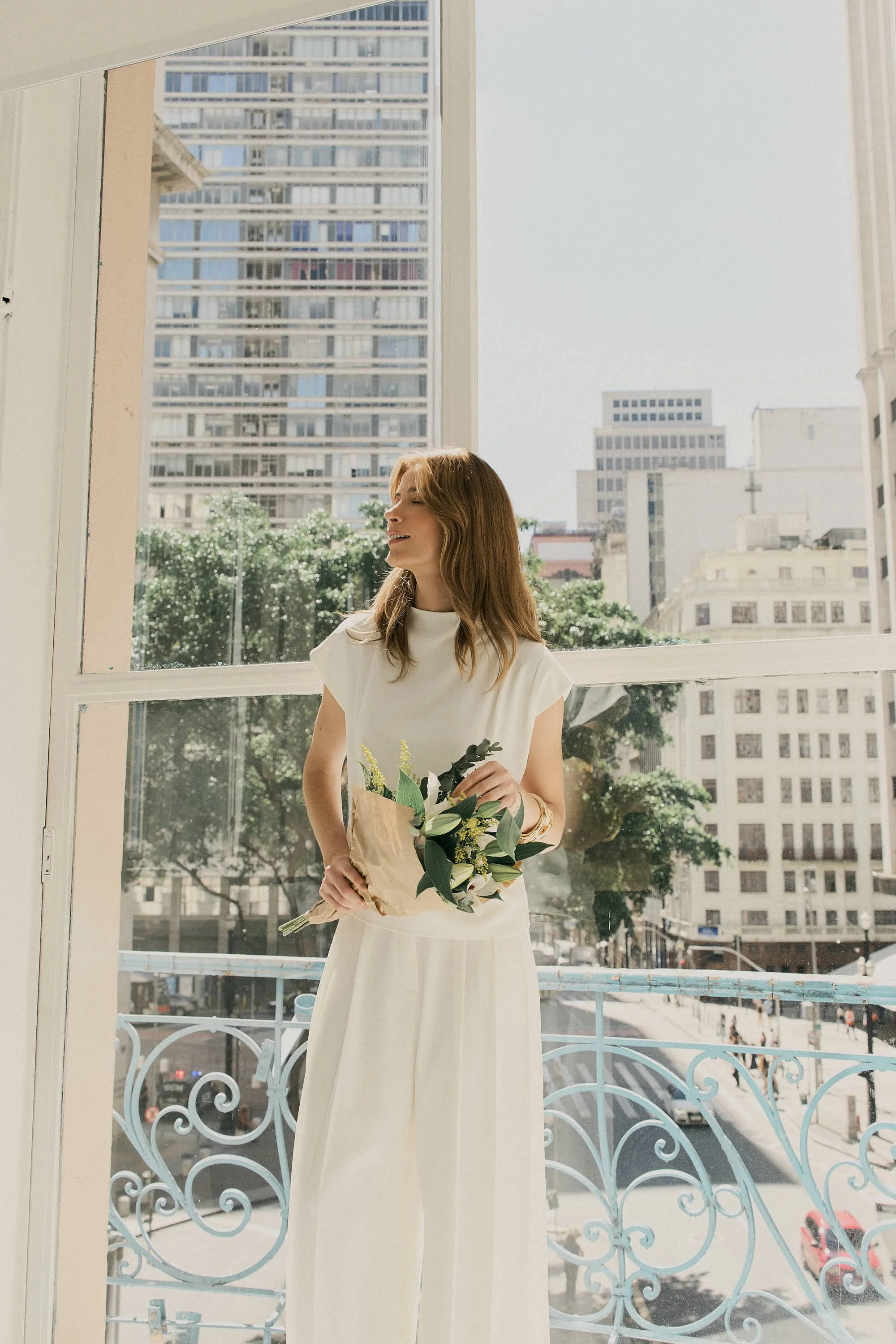 Mulher sorrindo segurando um buquê de flores, vestindo vestido branco, de pé perto de uma janela com grades decorativas, com vista para prédios na cidade ao fundo, durante o dia.