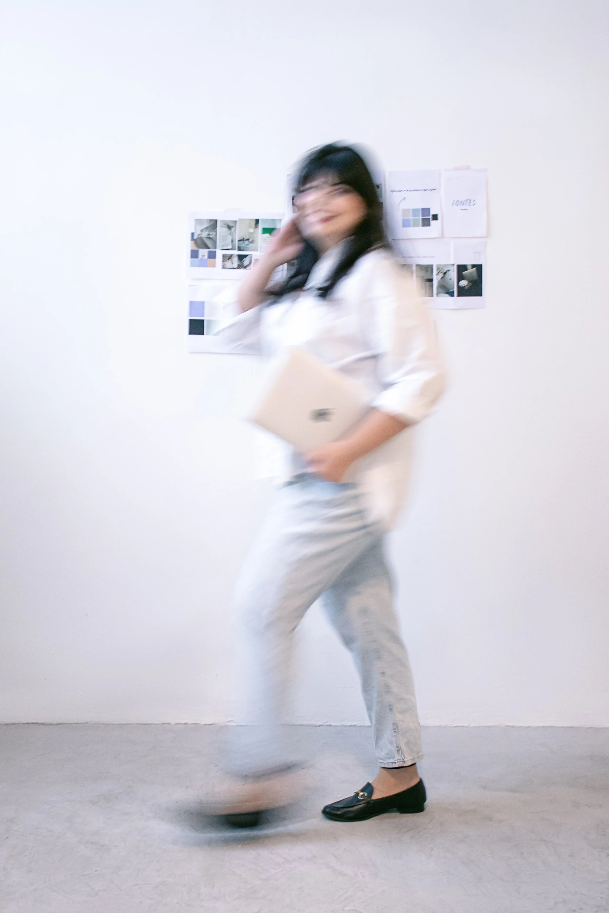 Pessoa com cabelos pretos, usando óculos, roupa branca e jeans, andando de lado em um ambiente branco, segurando um laptop e com um quadro de fotos na parede ao fundo.