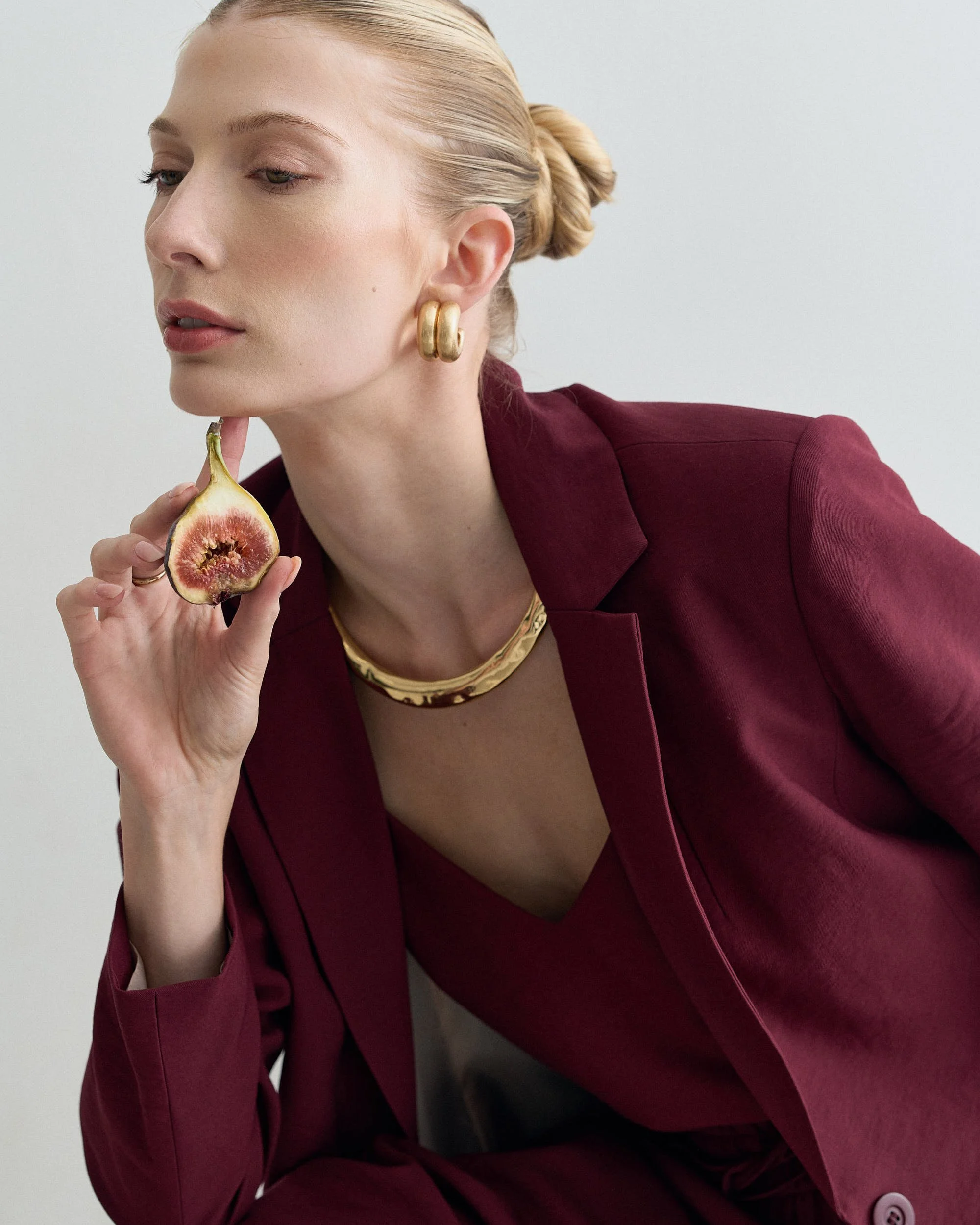 Mulher com cabelo preso, usando brincos grandes de argola dourada, blusa ou blazer vinho, usando colar dourado, segurando um figo cortado na mão, com fundo branco.