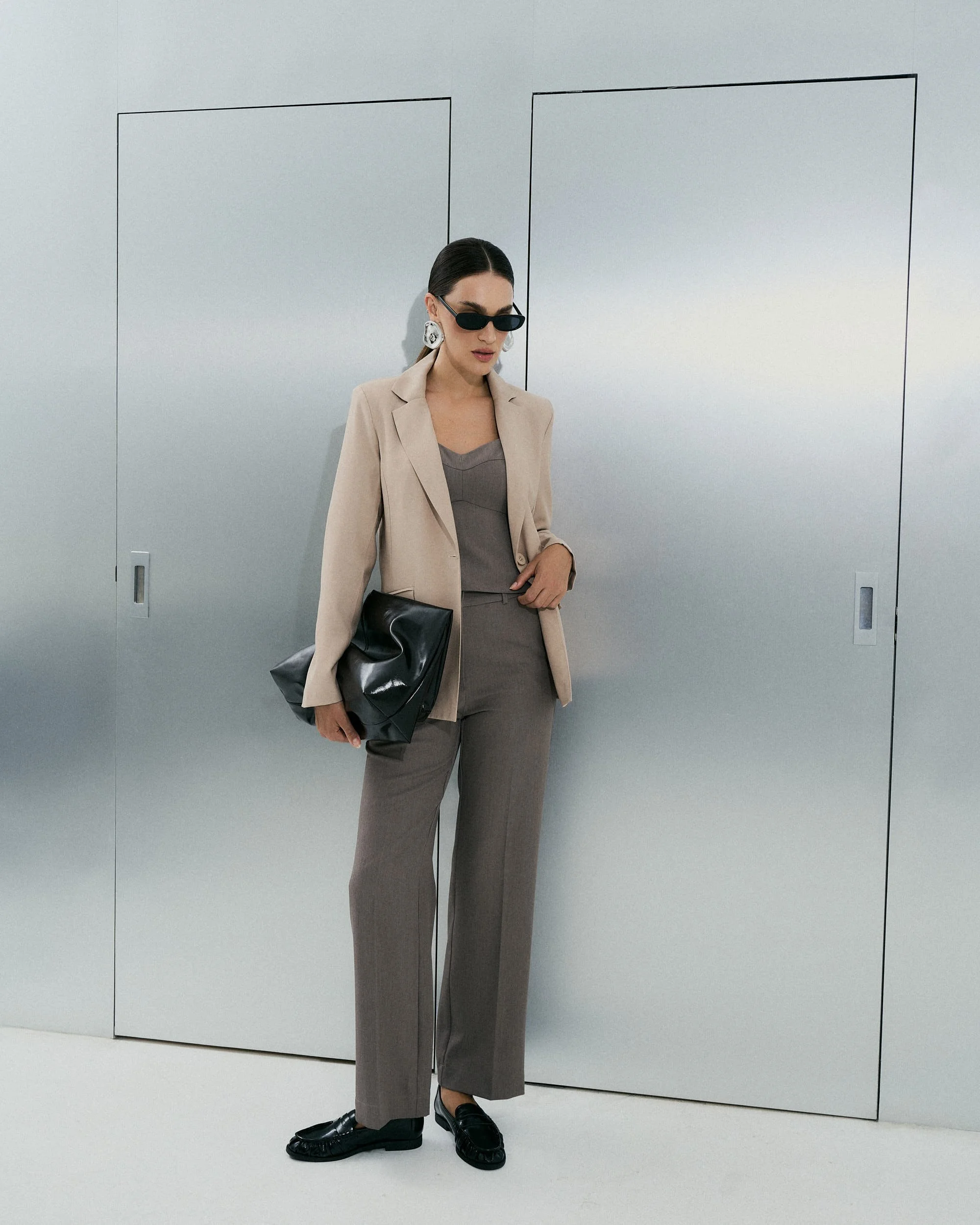 Mulher com cabelo escuro preso em um coque, usando óculos escuros de armação pequena, blazer bege, lenço cinza, calça cinza e sapatos pretos, segurando uma bolsa grande preta, em parede branca com linhas pretas.