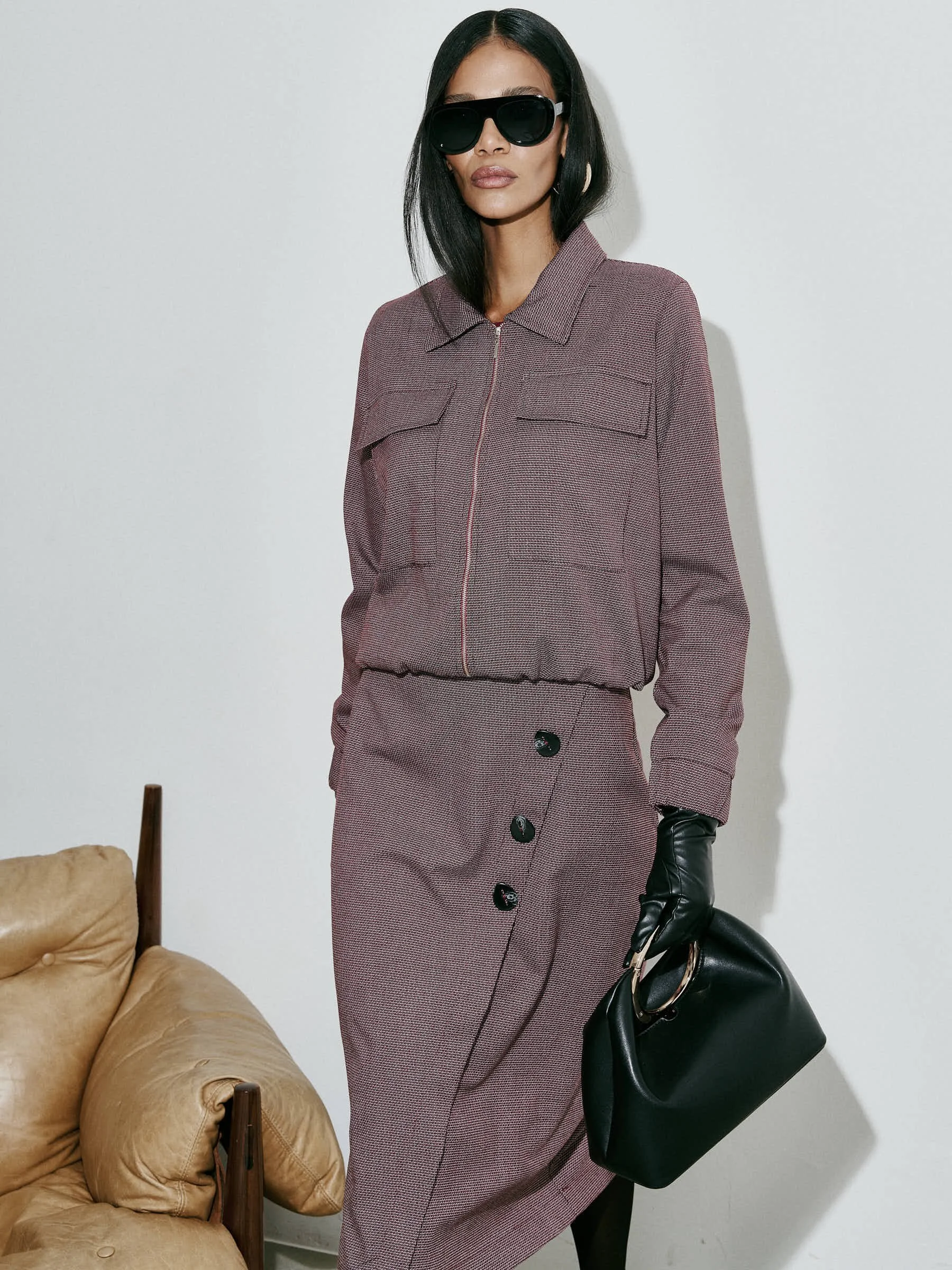 Mulher vestida com blazer e saia de tecido xadrez em tom de rosa, usando óculos escuros pretos e luvas pretas, segurando uma bolsa preta com alça de corrente.