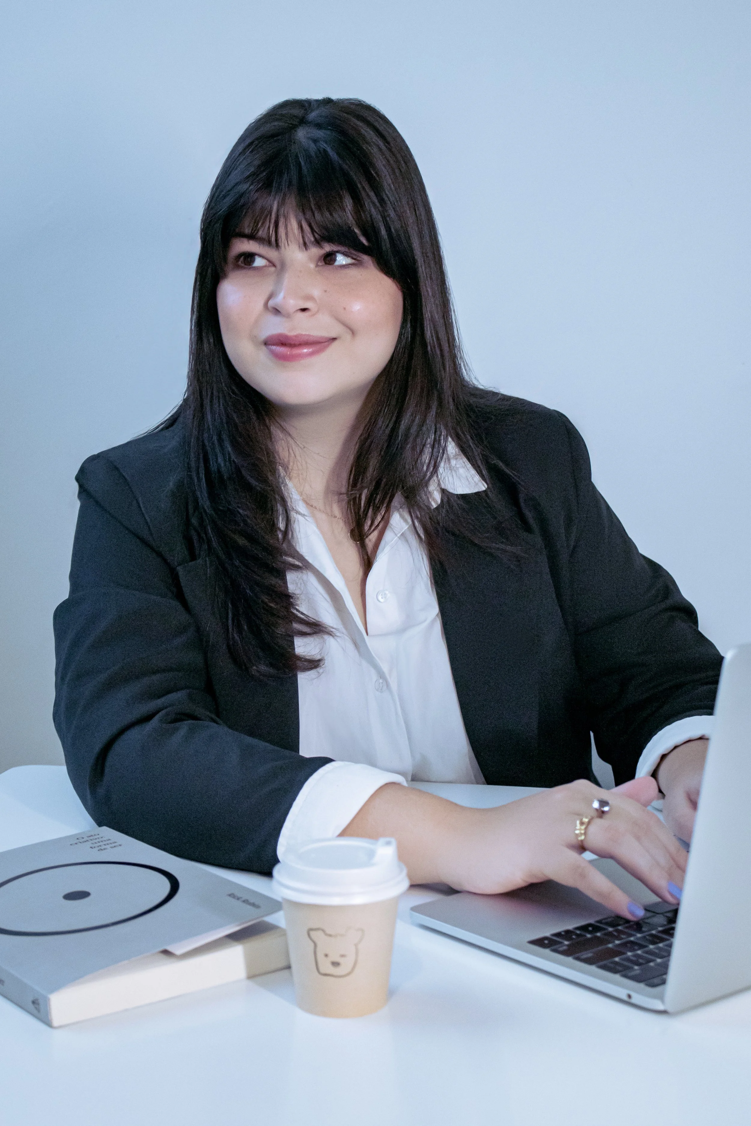 Mulher com cabelo preto, vestindo blazer preto e camisa branca, sentado em uma mesa de escritório, usando um laptop, com uma xícara de café ao lado e uma mesa com livros e material de escritório.