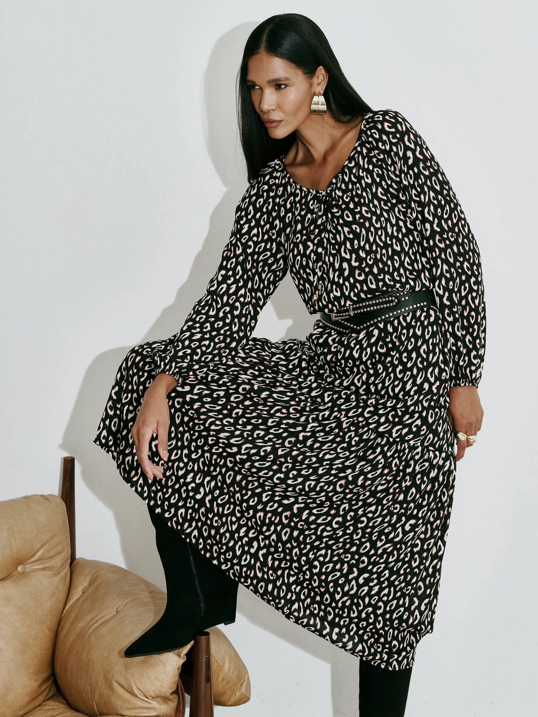 Mulher usando vestido preto com estampa de onça, com acessórios dourados, apoiada em uma peça de mobiliário com uma perna apoiada nela, contra fundo branco.