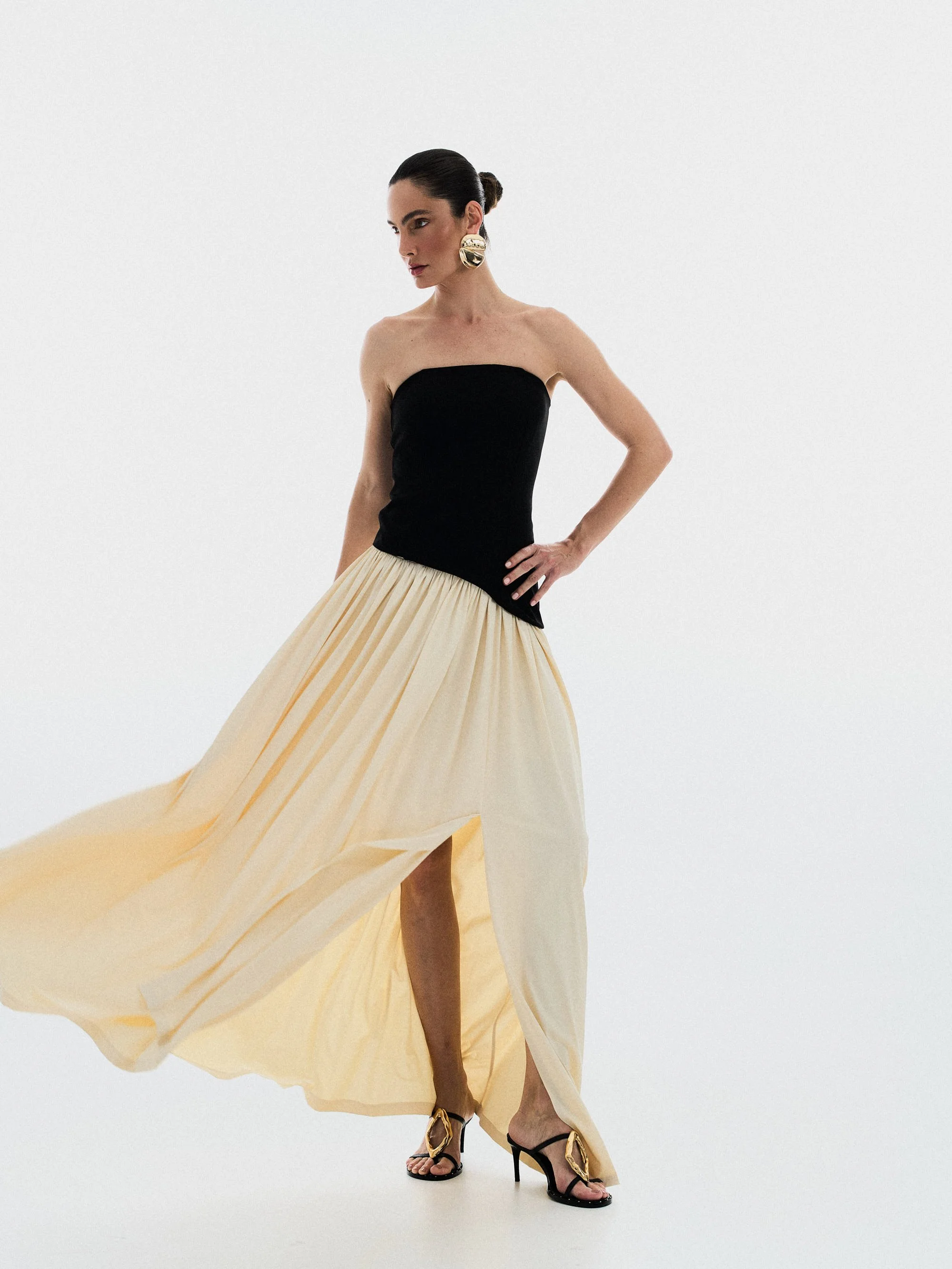 Mulher de vestido preto e saia longa amarela, usando bigode dourado e brincos grandes dourados, com cabelo preso para trás, usando sandálias pretas com detalhes dourados, posando com uma mão na cintura em um fundo branco.