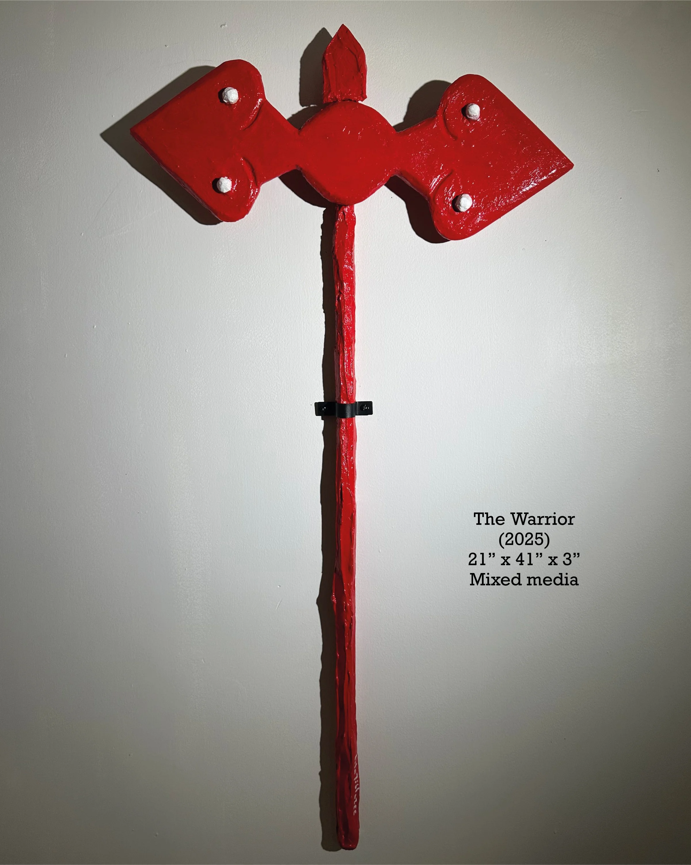 Red abstract sculpture resembling a warrior with a pointed top and a long, vertical tail in the center, mounted on a light-colored wall.