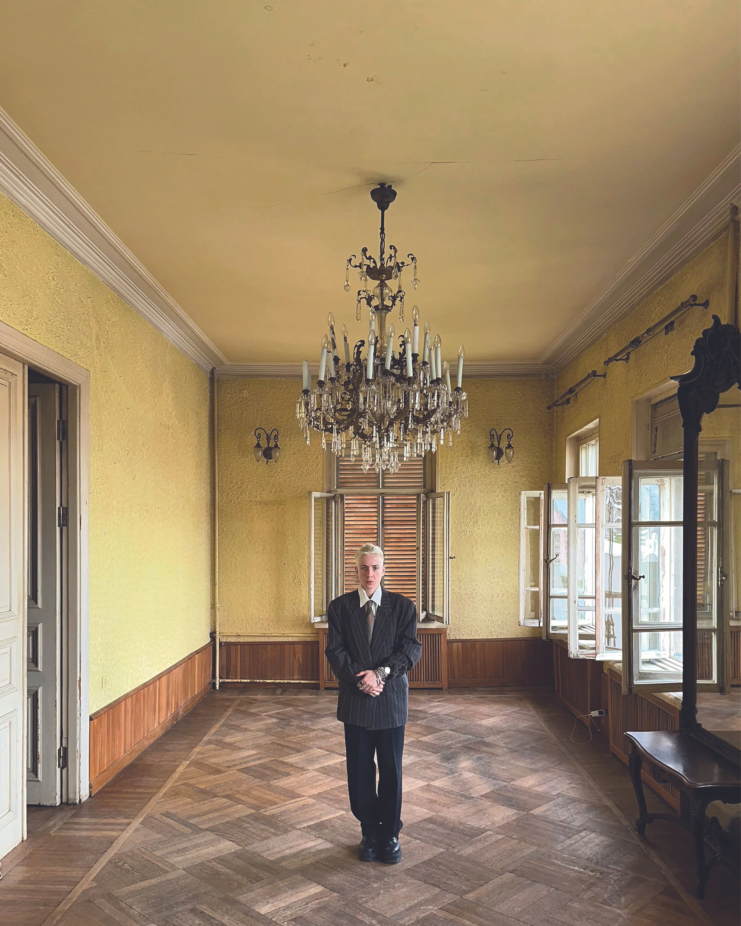 A woman standing in a room with yellow walls, large windows, and a chandelier, dressed in a dark pinstripe suit with a white shirt, in a formal setting.