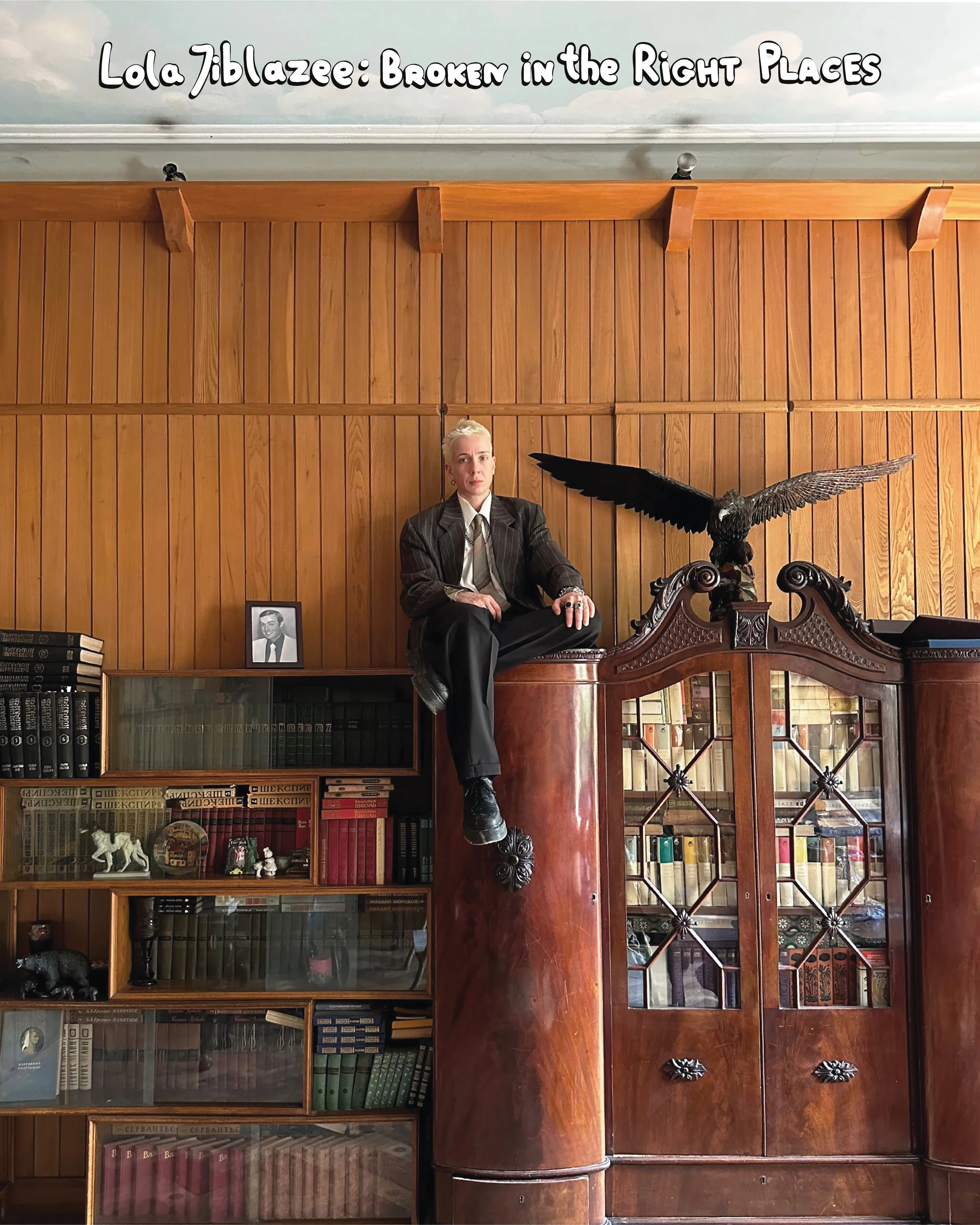 A woman with blond hair, dressed in a pinstripe suit, sits on a large vintage wooden desk in a room with wooden paneled walls. There is a black and white photograph of a man, a bookshelf filled with books, and decorative items including a black pig f