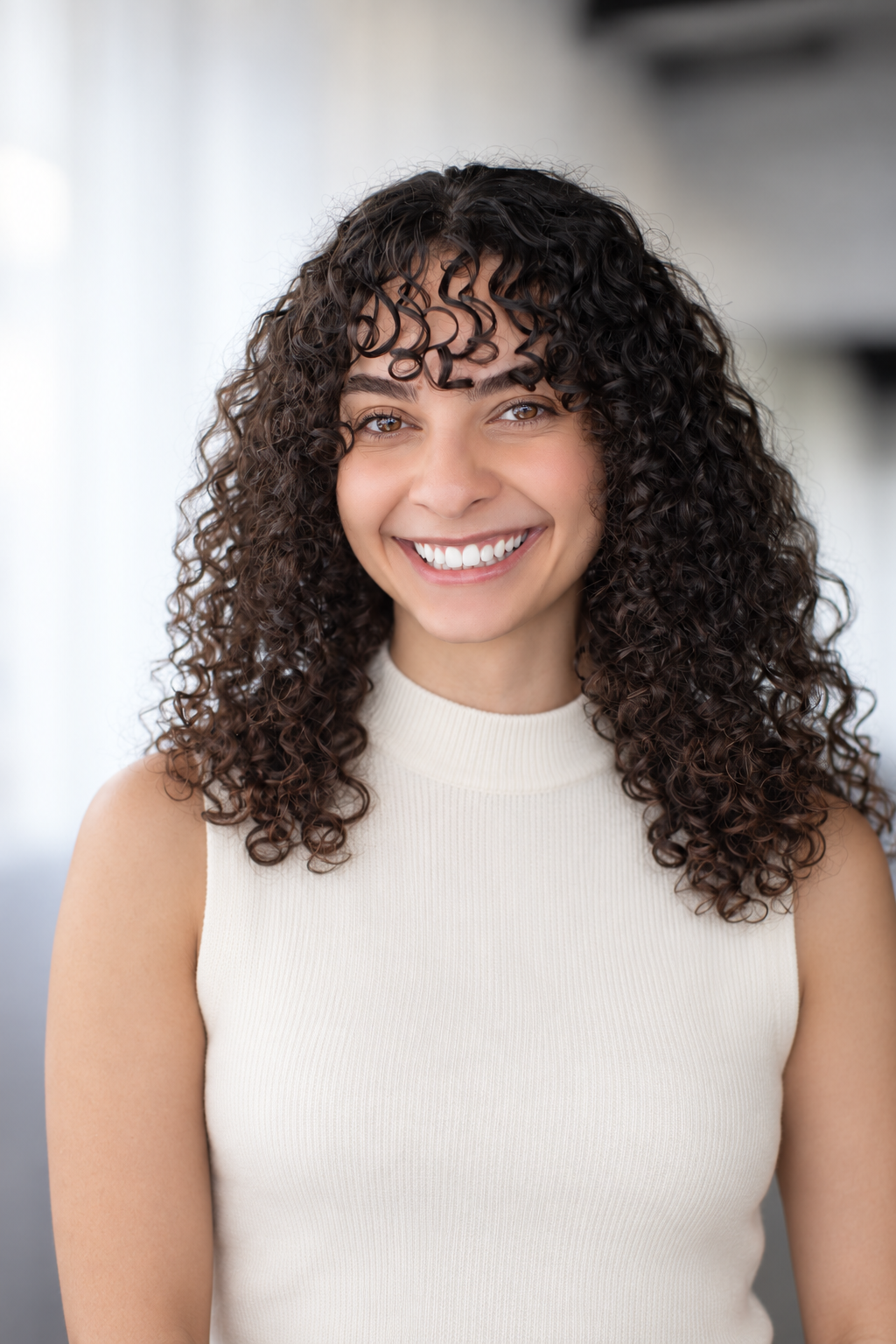A woman with curly dark hair smiling at the camera, wearing a sleeveless cream top, indoors with a bright background.