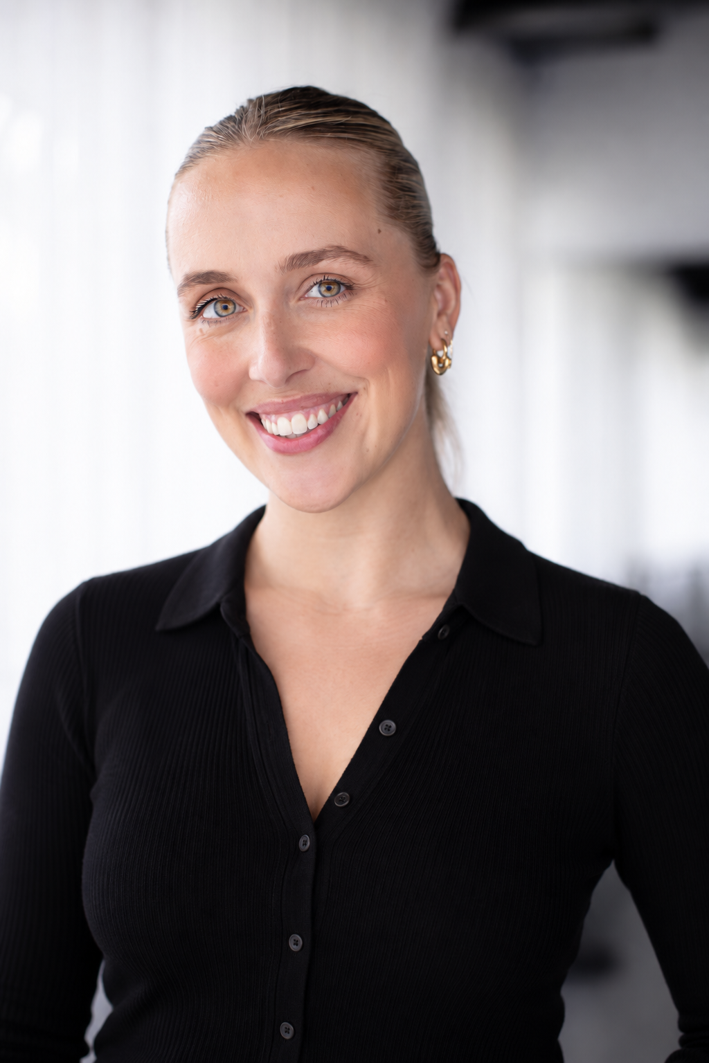 A young woman with blonde hair tied back, smiling, wearing a black button-up shirt and gold earrings indoors.