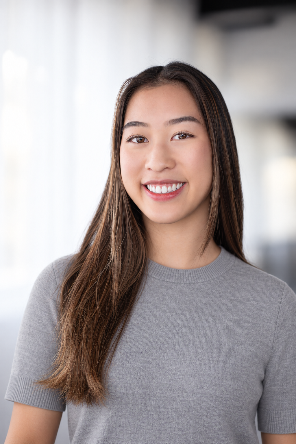 Portrait of a young woman with long brown hair smiling, wearing a light gray t-shirt, standing indoors with bright, natural lighting.
