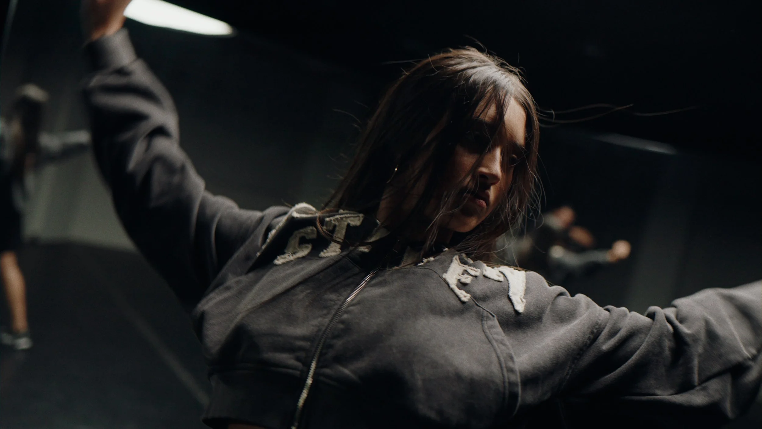 A woman with long, messy hair and a serious expression wearing a dark hoodie, with a dark background.