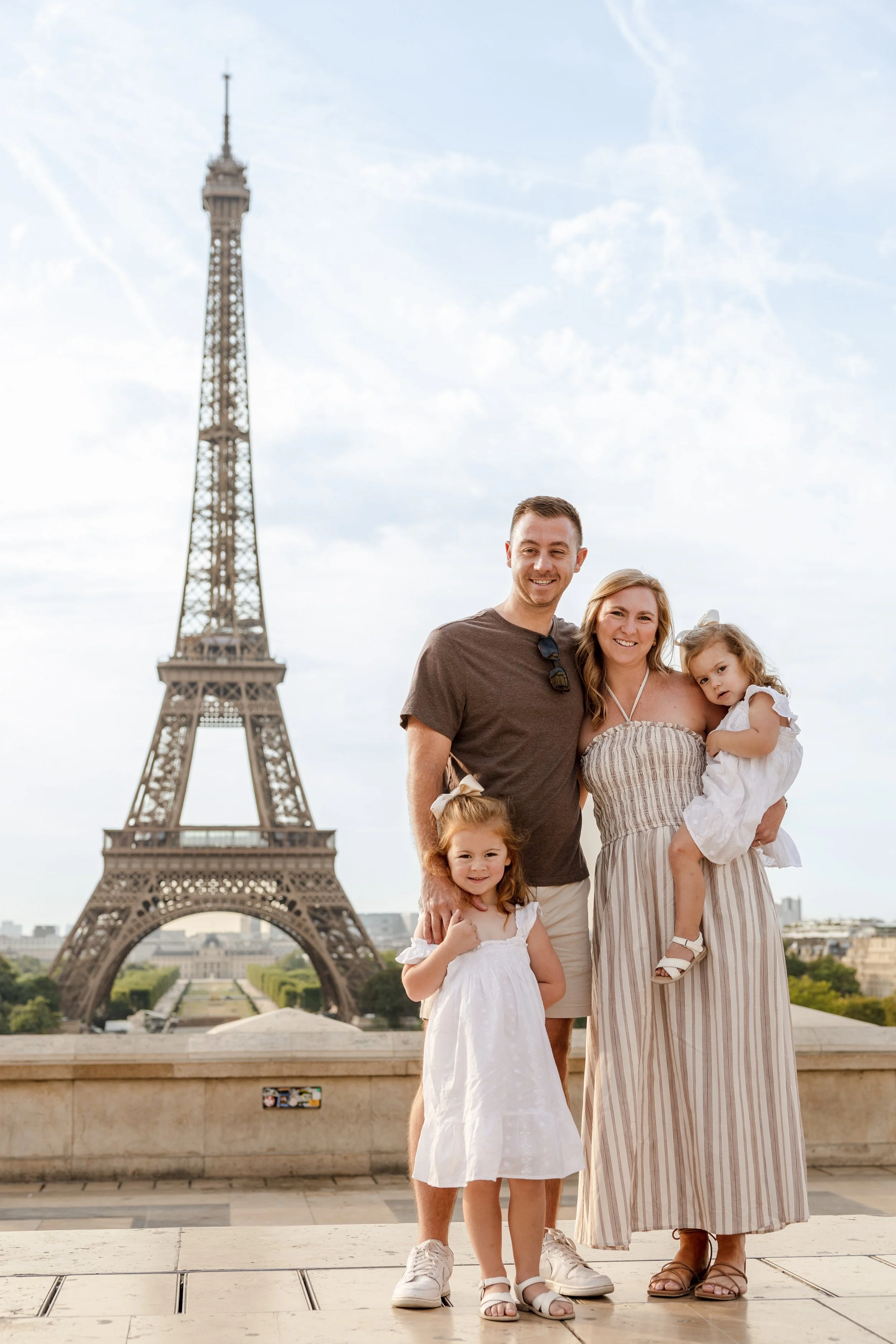 Family of four posing in front of the Eiffel Tower in Paris, France. The father, mother, and two young daughters are smiling at the camera on a sunny day.