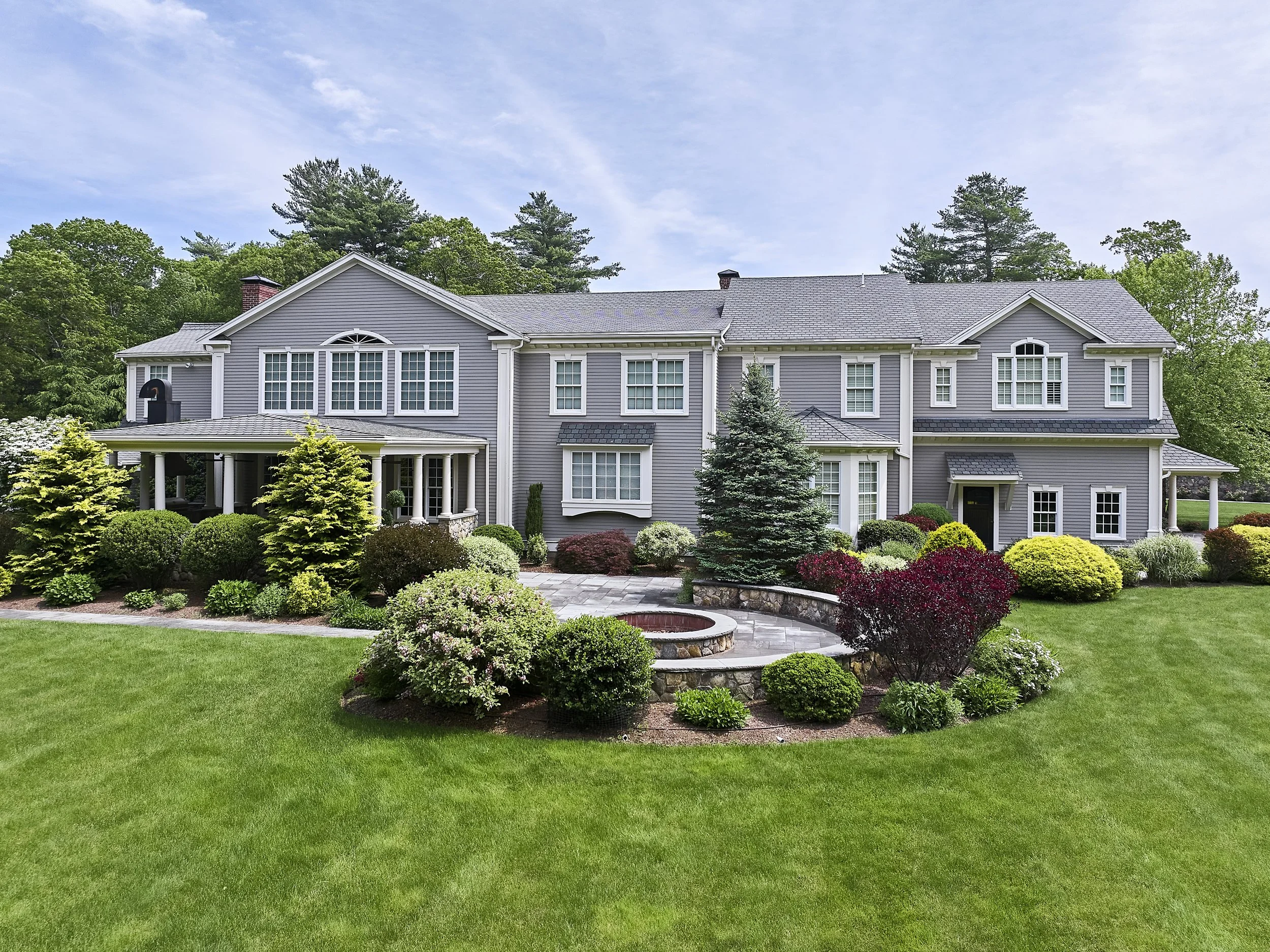 A large two-story house with gray siding and white trim, surrounded by a well-maintained garden with green shrubs, colorful bushes, and a paved walkway, set against a backdrop of tall trees and a partly cloudy sky.