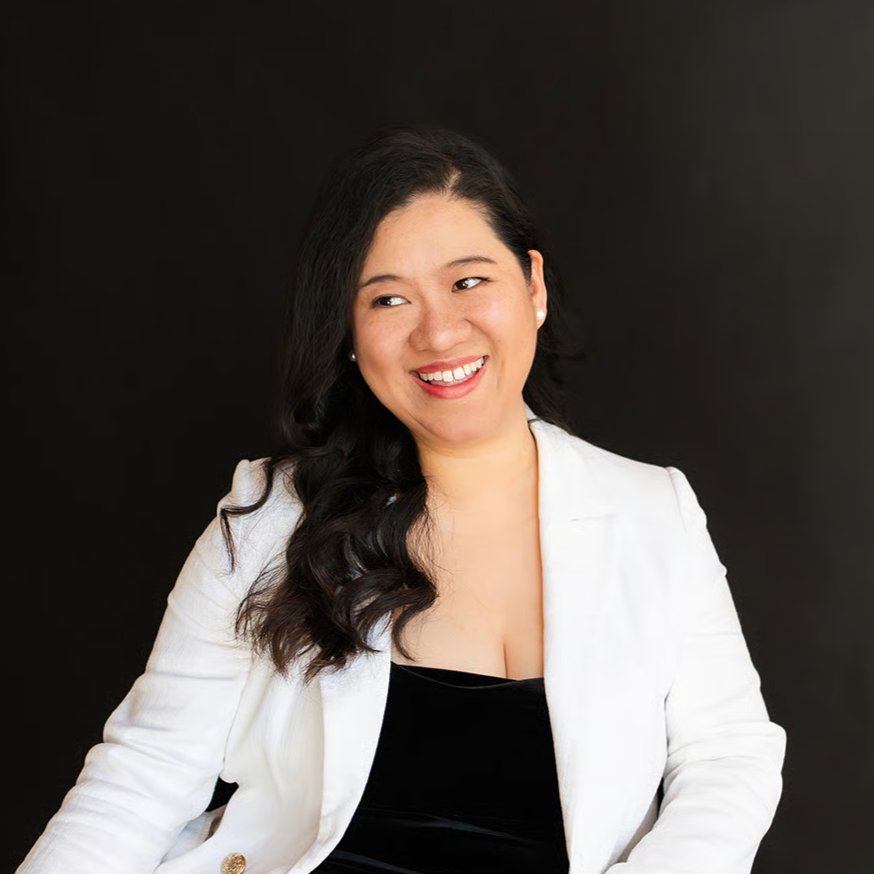 A woman with long dark hair, smiling, wearing a white blazer and black top, against a black background.