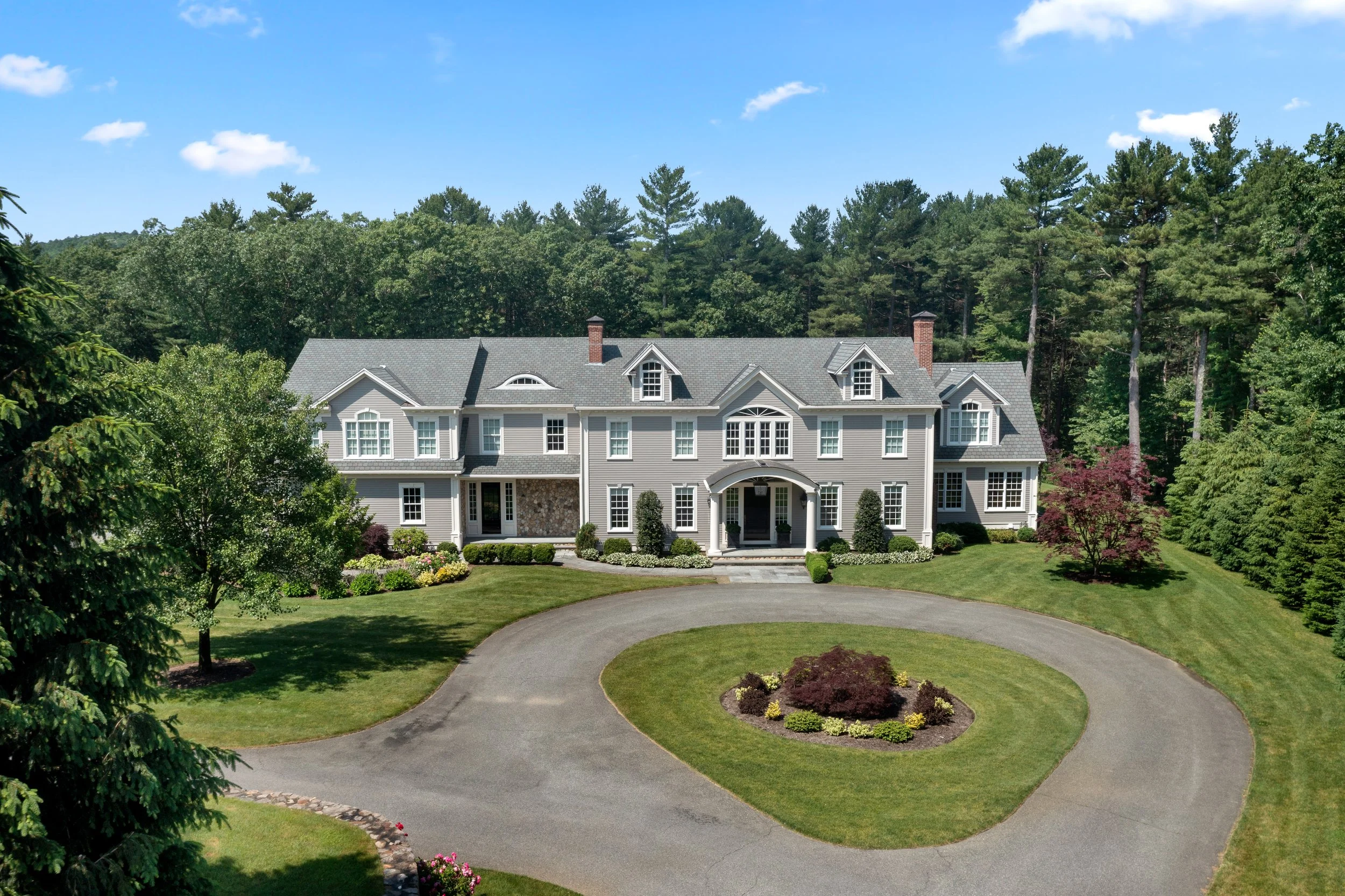 Large two-story house with gray siding and multiple gables, set against a lush green forest and blue sky, surrounded by a circular driveway and landscaped yard.