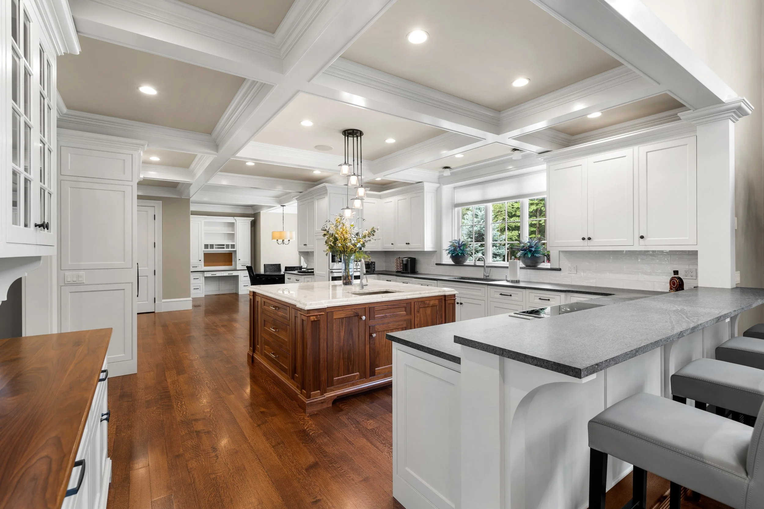 Bright modern kitchen with white cabinets, granite countertops, a central wooden island with drawers, and hardwood floors. Large windows provide natural light, and there are decorative plants on the windowsill. Recessed lighting and a hanging light f