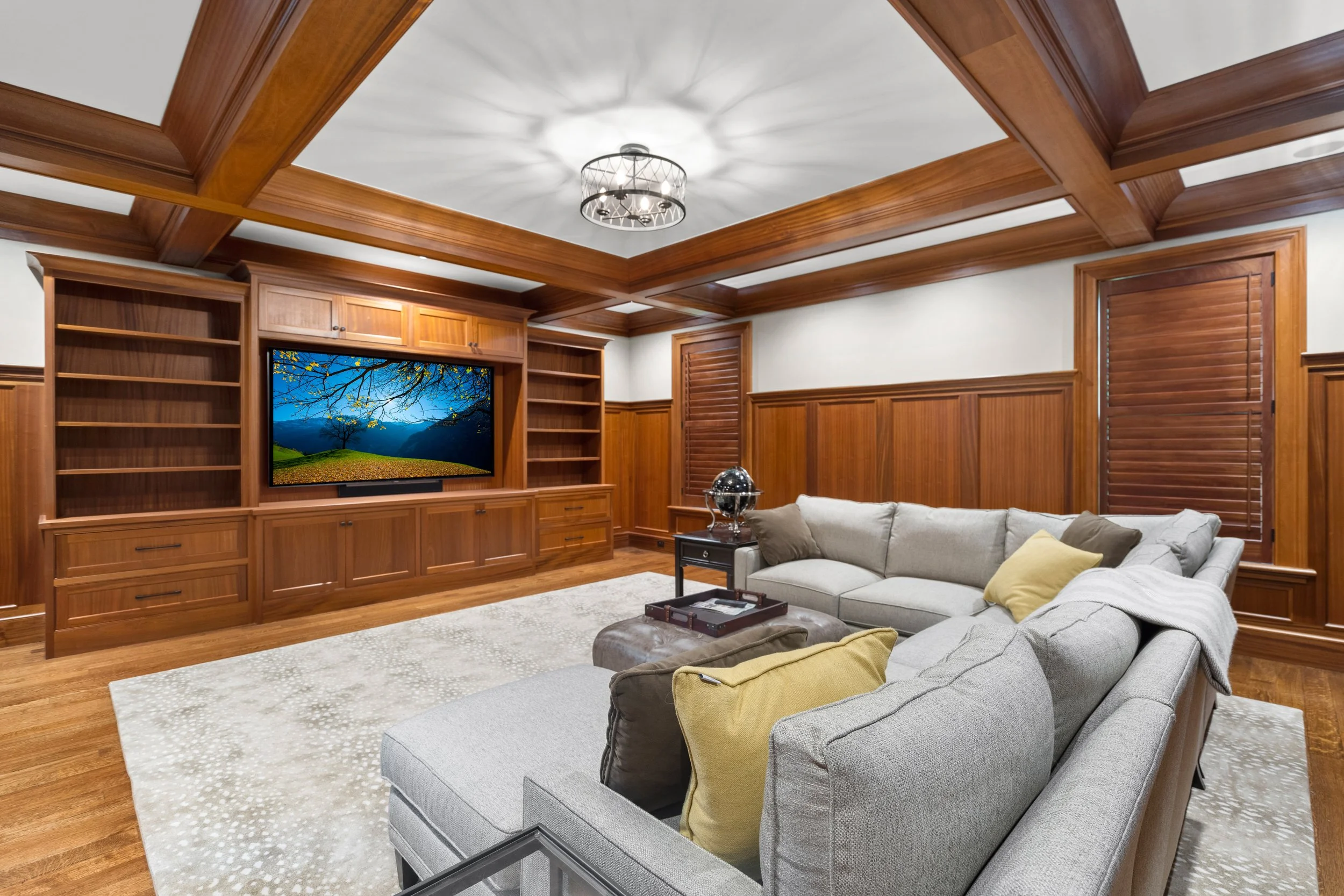 Living room with wood-paneled walls and ceiling, a large beige sofa with pillows, a TV showing a landscape, and a plush white rug.