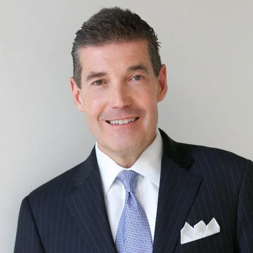 A smiling man wearing a dark pinstripe suit, white dress shirt, and light blue tie, standing against a plain light-colored background.