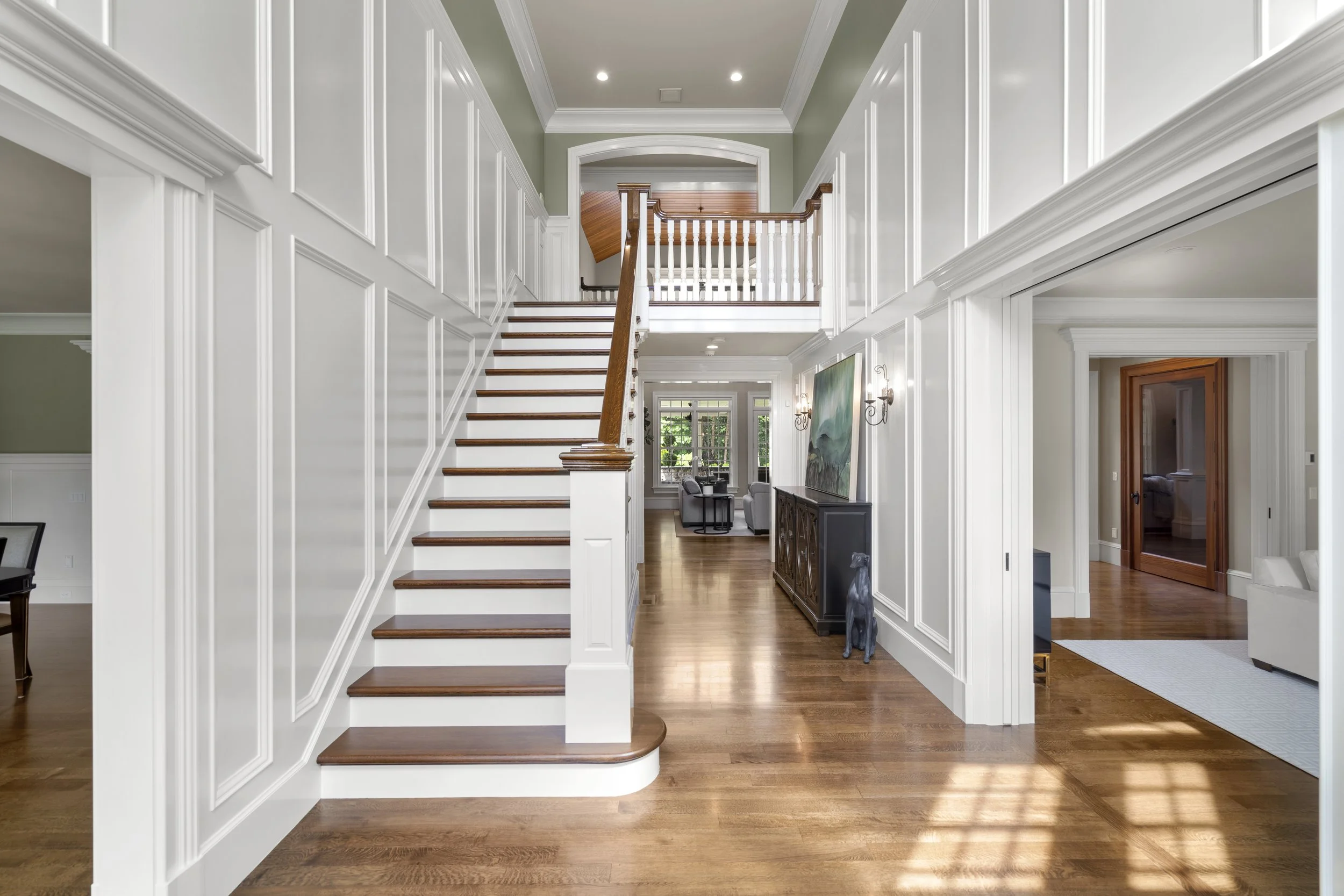 Interior view of a spacious house entryway with hardwood floors, white wainscoting, staircase, and a sitting area with chairs, along with artwork and wall sconces.