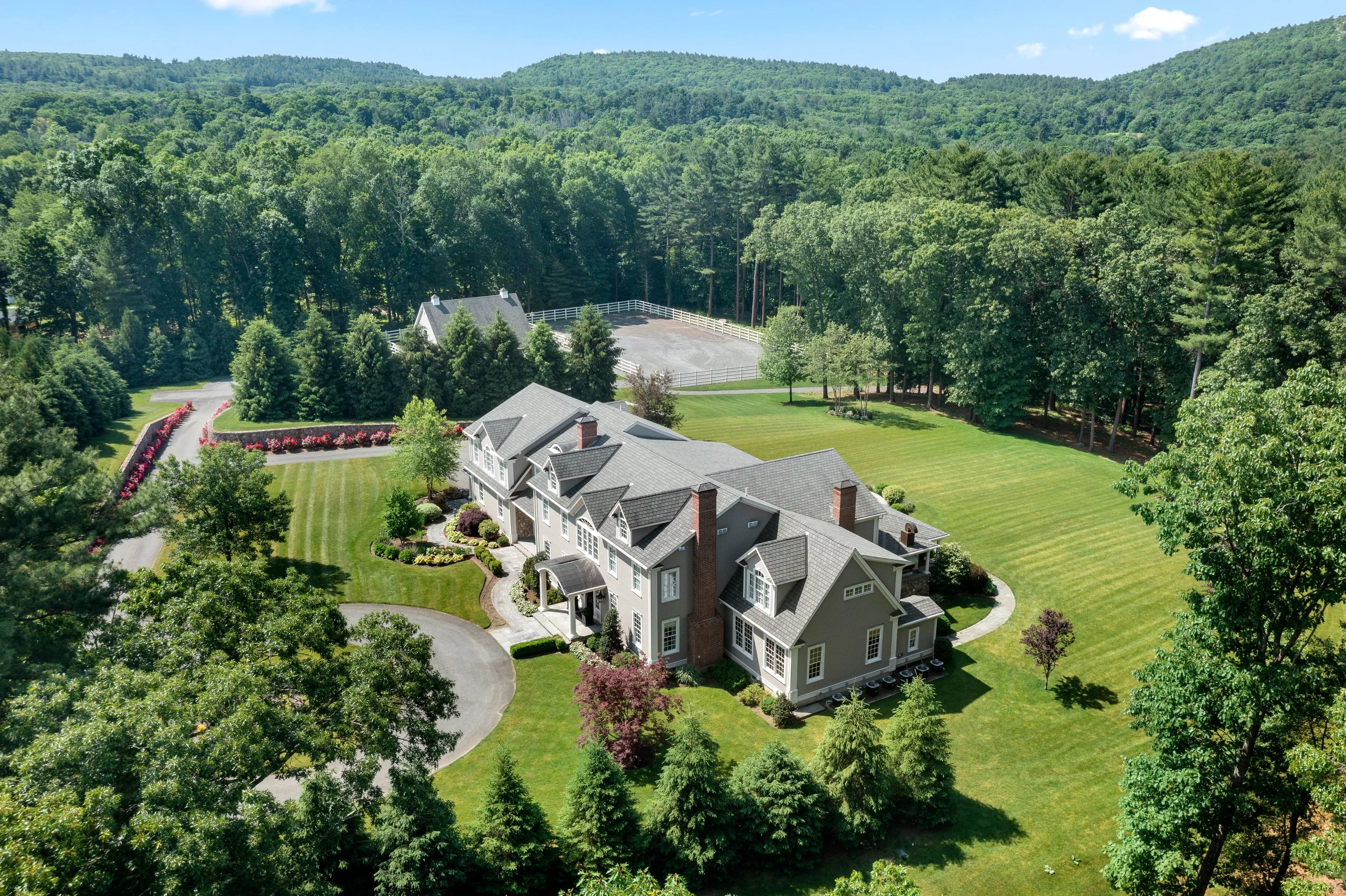 A large house with a gray roof and brick chimneys surrounded by green trees and a well-manicured lawn. There is a winding driveway with a flower bed along the side, and a forested hillside in the background.