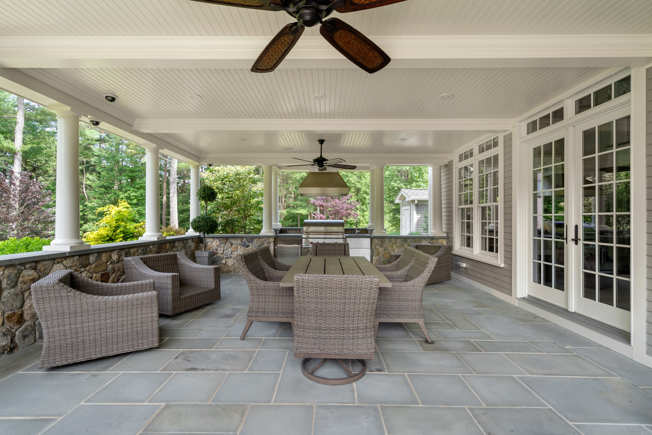 Covered outdoor patio with a dining area, wicker chairs, a rectangular table, ceiling fans, large windows, a stone wall, and lush greenery in the background.