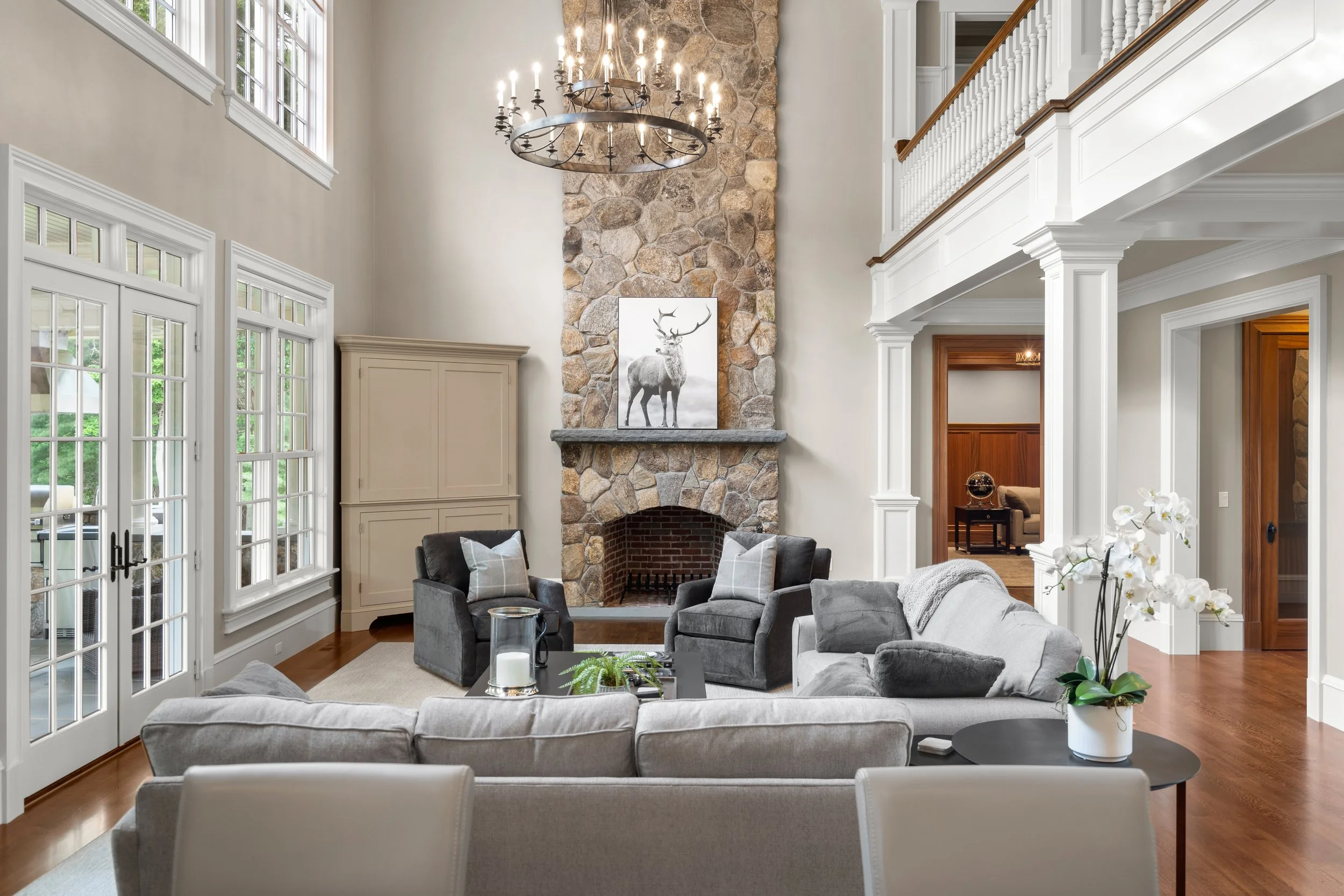 A spacious living room with large windows, a stone fireplace with a deer painting above it, and comfortable seating including sofas and armchairs. There's a chandelier hanging from the ceiling and a view of an outdoor patio.