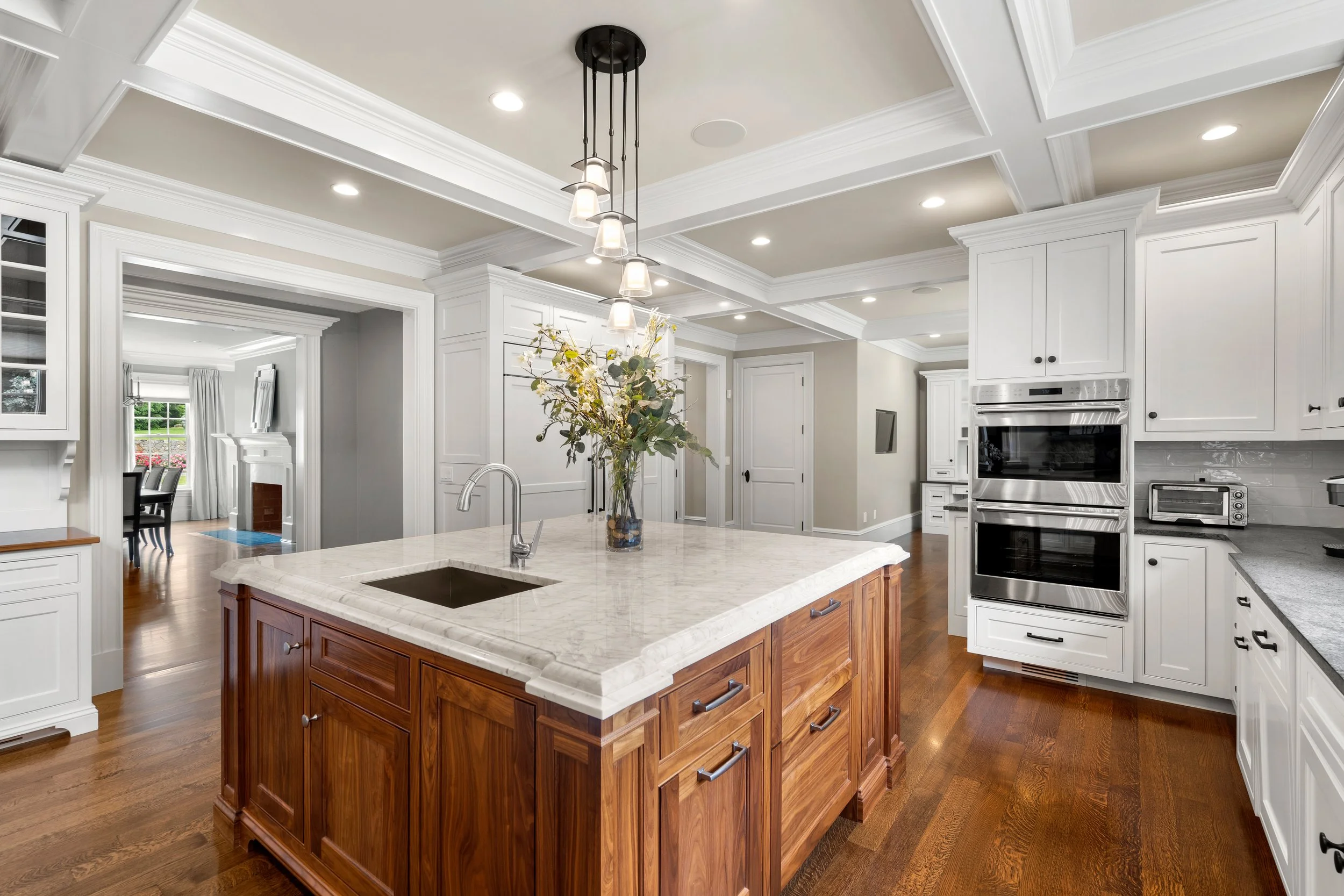 A modern kitchen with white cabinets, a large marble island with a wooden base, a vase with flowers, and built-in stainless steel appliances, including a double oven.