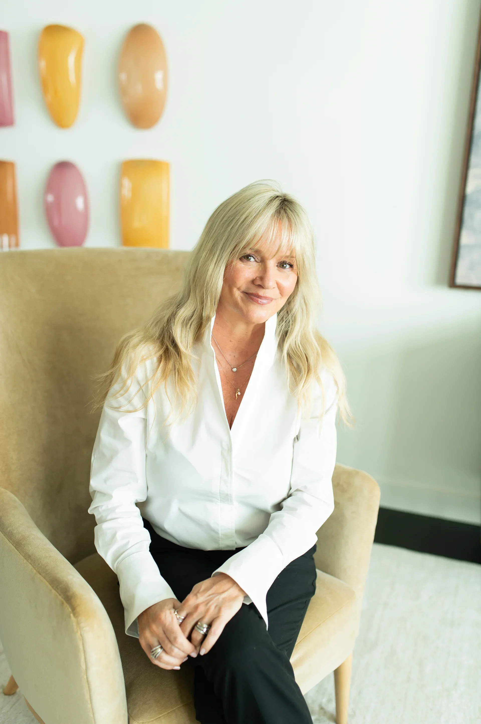 A woman with long blond hair, wearing a white shirt, sitting on a beige armchair in front of a white wall with colorful ceramic wall art. She is smiling and has her hands resting on her lap.
