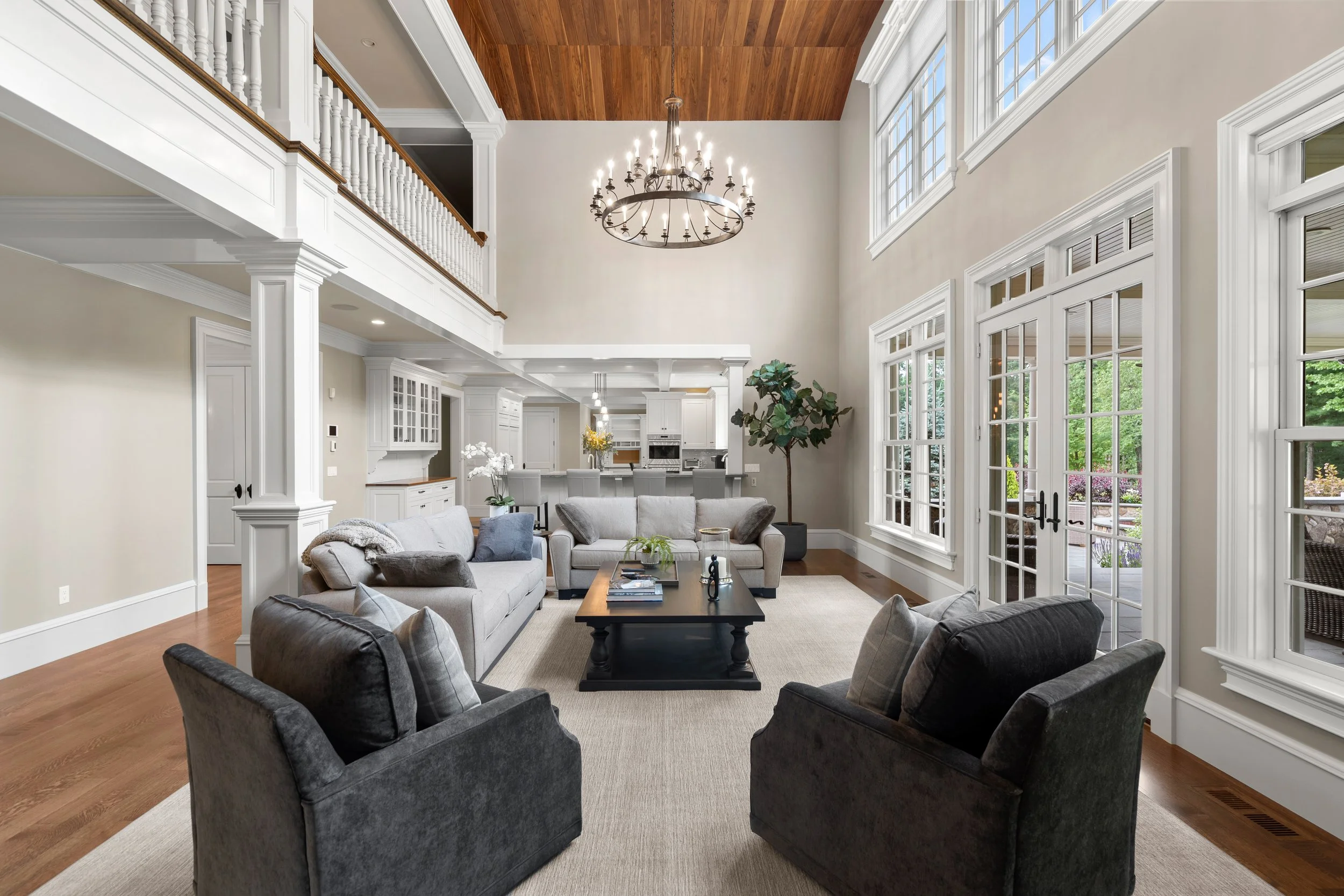 A spacious living room with beige walls, large windows, white trim, and a wooden ceiling. The room features multiple gray and black armchairs around a black coffee table, a white sofa, and decorative plants. There's a chandelier hanging from the ceiling and a view of a kitchen area in the background.