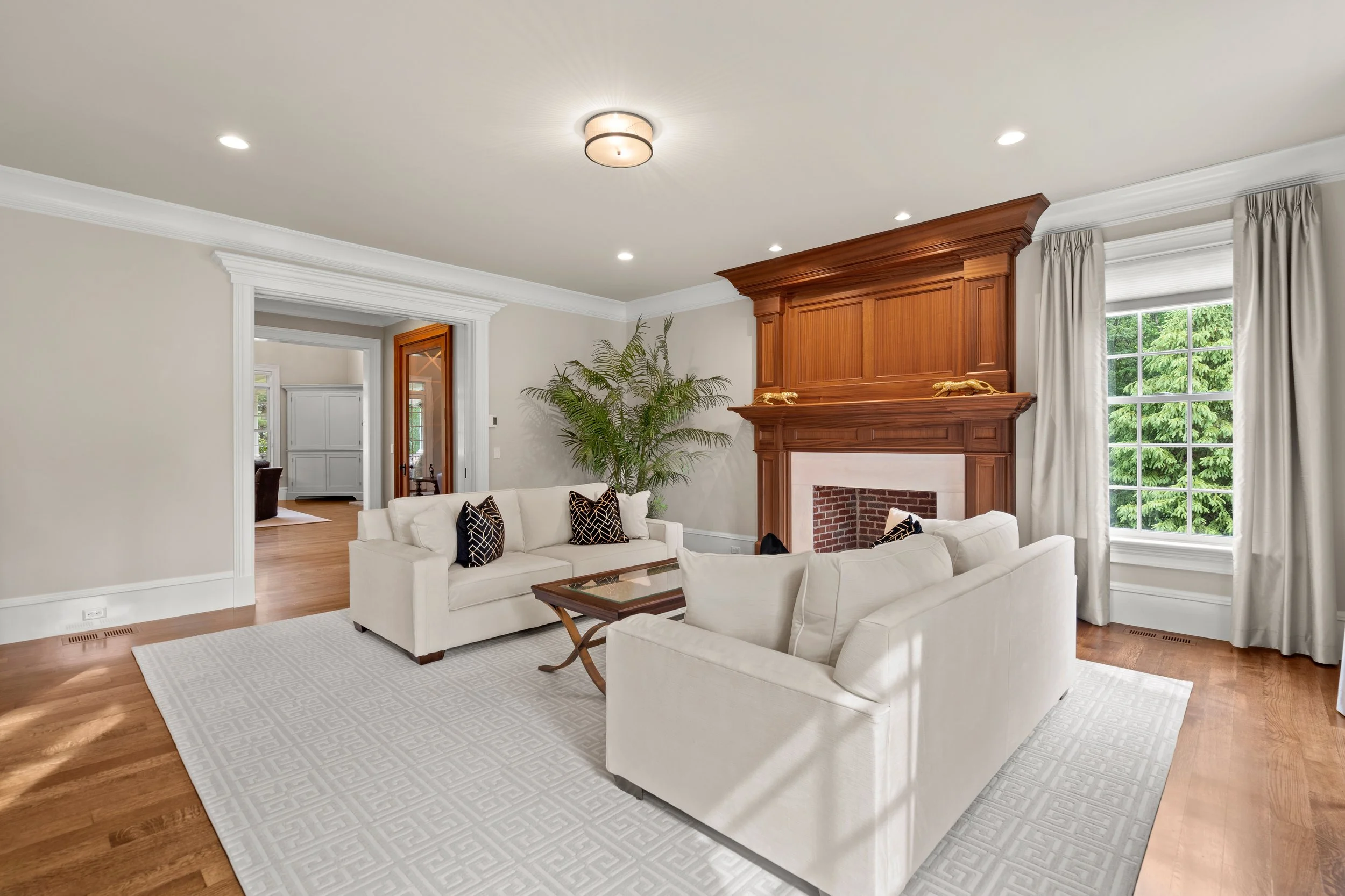 Living room with white sofas, black and gold pillows, wooden fireplace mantel with gold animal decorations, large window with curtains, green trees outside, hardwood floor, ceiling light, and recessed lighting.