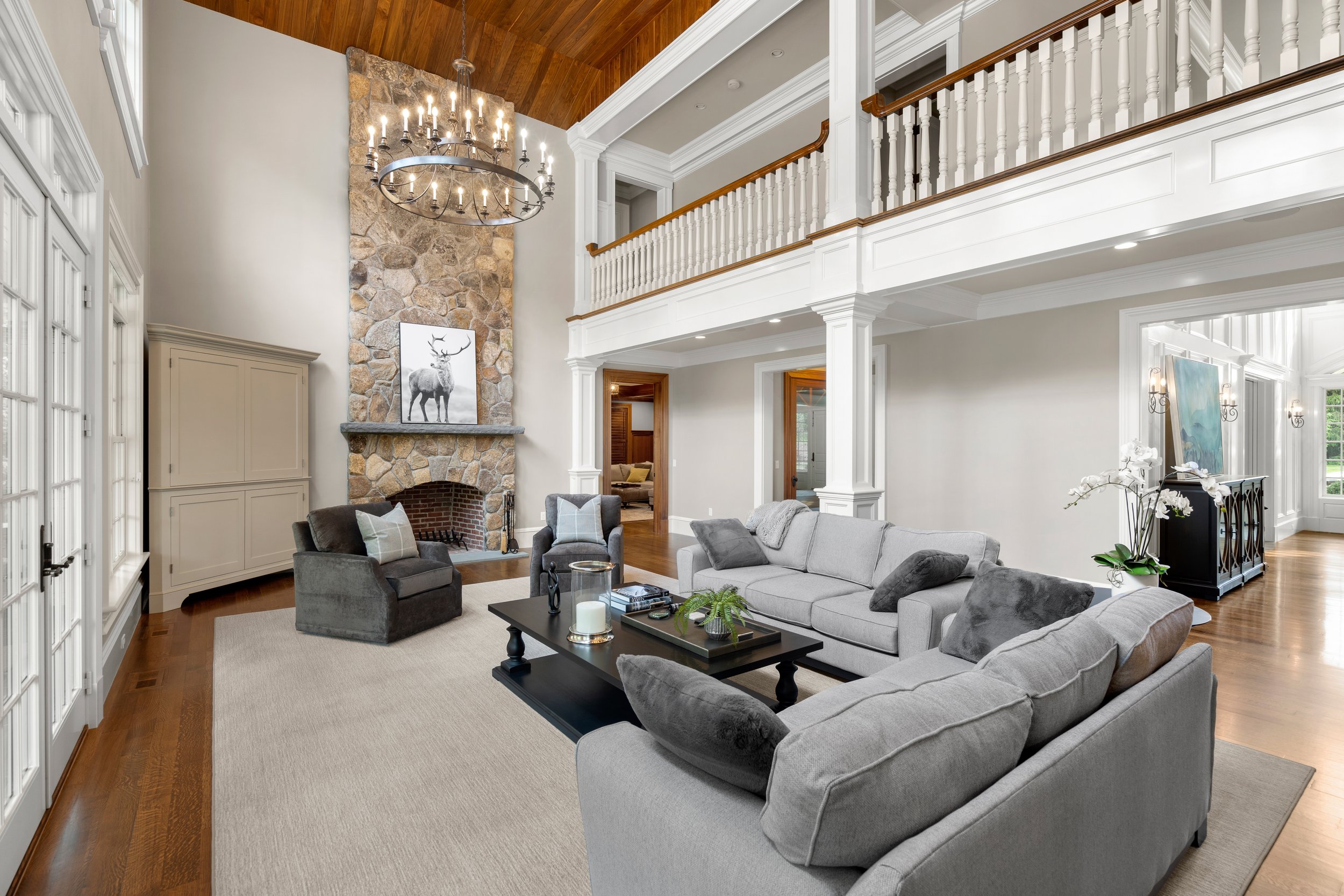 Living room with high vaulted wooden ceiling, stone fireplace with a deer artwork on the mantle, chandelier, multiple windows, and gray sofas.