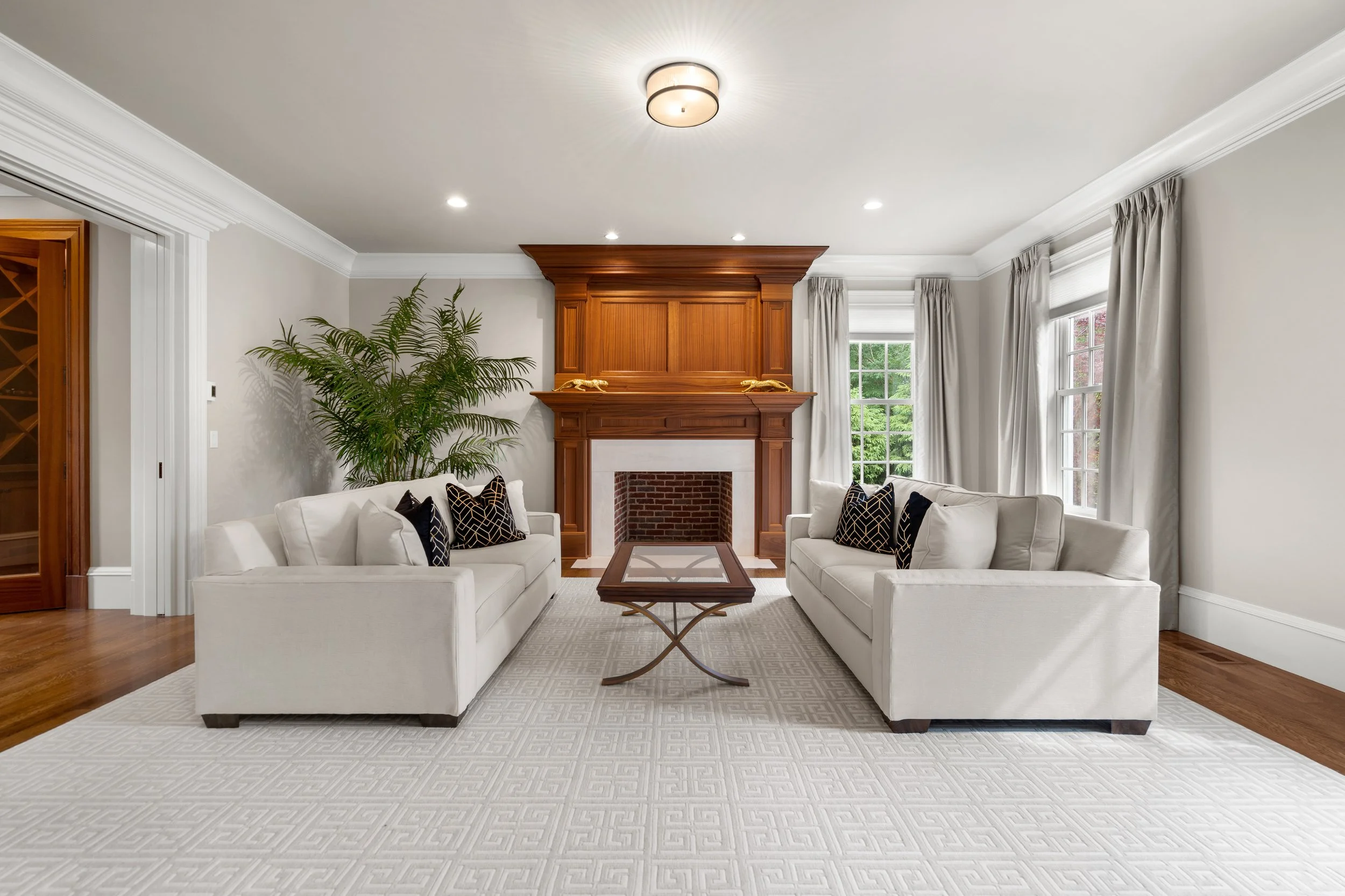 Living room with white sofas, black patterned pillows, a wooden coffee table, a large green plant, a wooden fireplace mantel, large windows with white curtains, and a white patterned area rug.