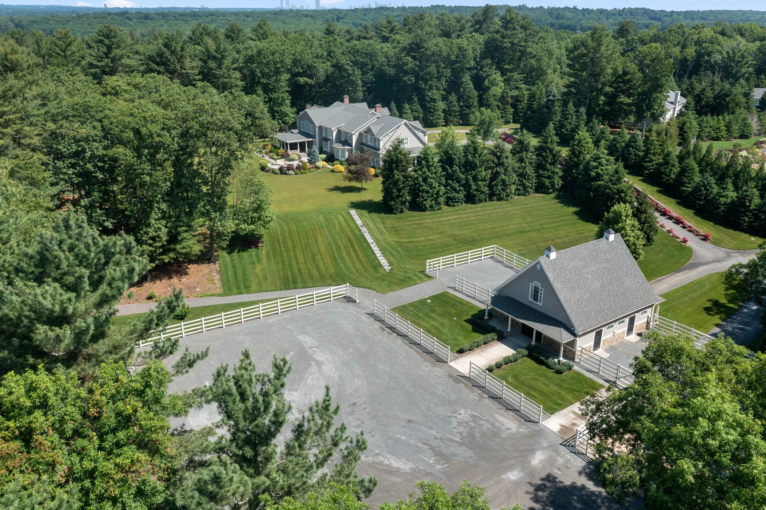A large house surrounded by a well-maintained lawn, trees, and a white fence, with a gravel driveway and a separate garage structure, in a green, wooded area with distant city skyline in the background.