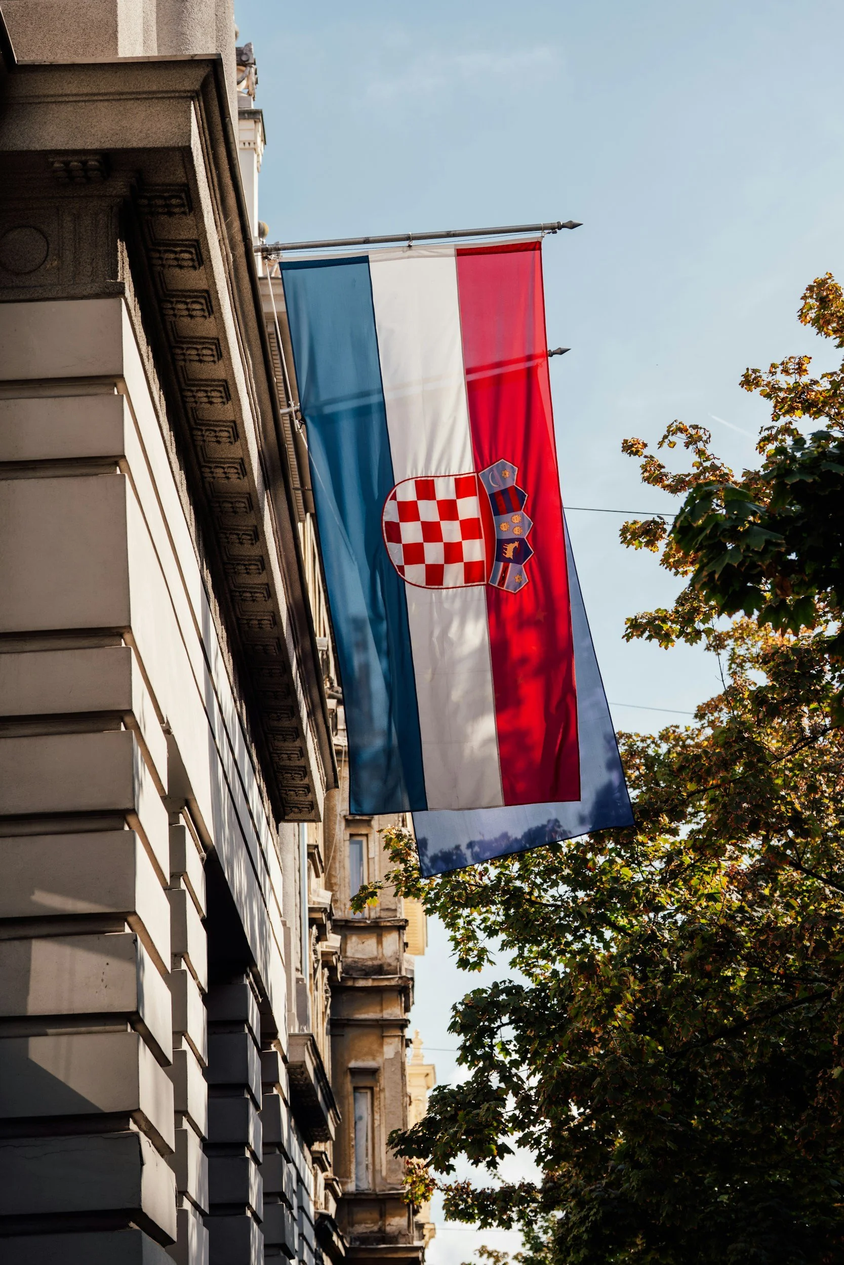 A Croatian flag hanging from a building with trees and other buildings in the background on a clear day.