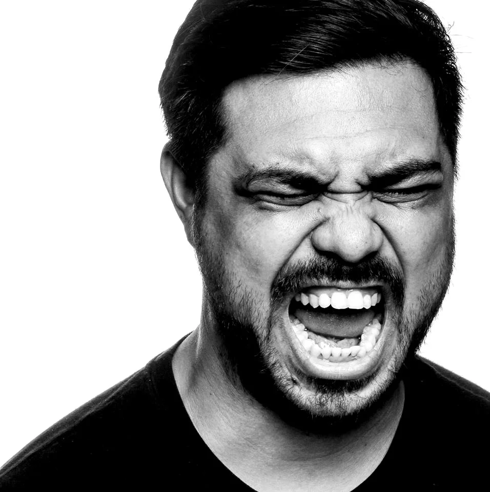 A black and white close-up photo of a man with a beard and short hair, mid-shout or scream, with an intense expression.