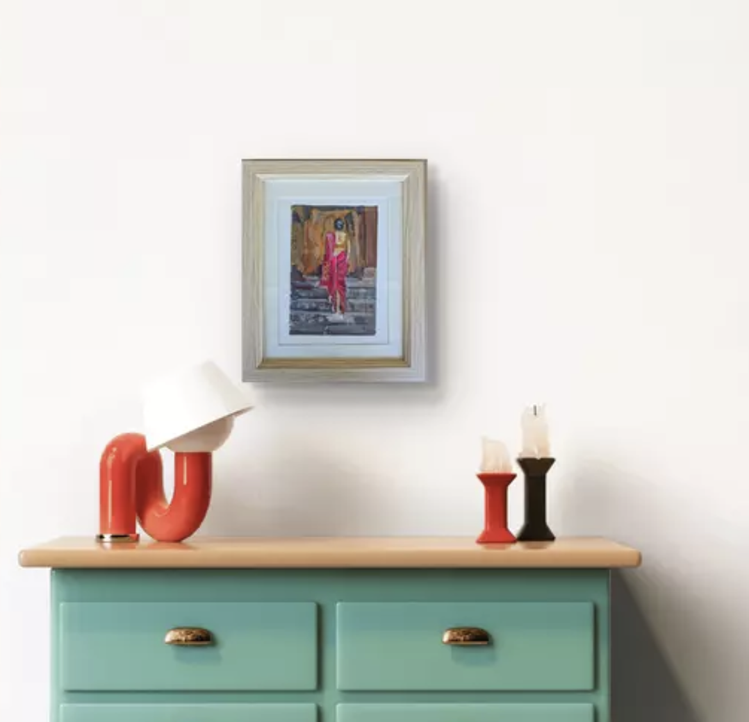 A mint green dresser with three drawers, topped with a modern table lamp with a white shade and curved red base, and two candle holders, one red and one black, with white candles. Above, a framed colorful painting of a woman in a pink  standing on stairs on a white wall.