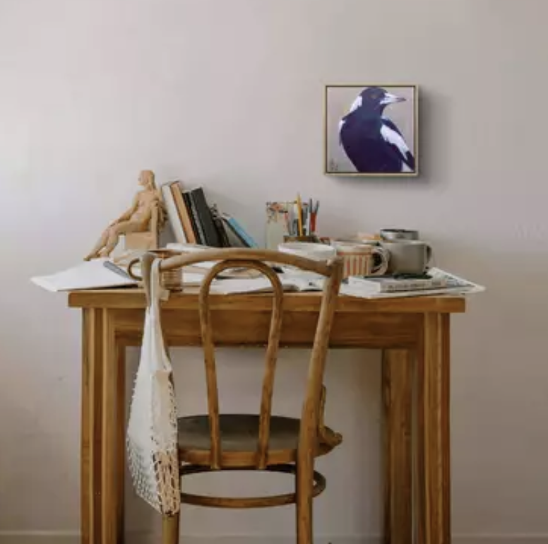A wooden desk with books, stationery, and a small wooden doll on top. A woven chair is in front of the desk. A framed picture of a magpie hangs on the wall behind the desk.