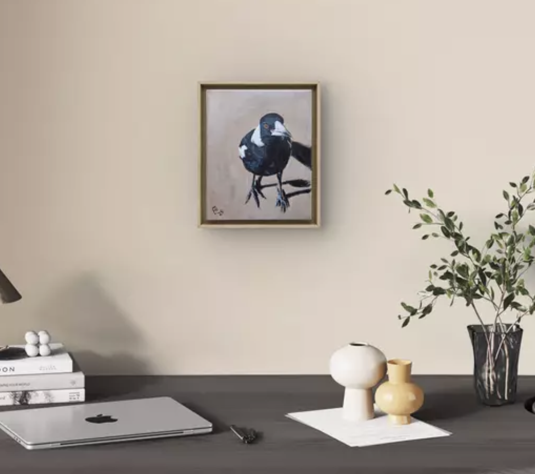 A framed painting of an Australian Magpie on a beige wall, a desk with a closed laptop, books, decorative vases, and a potted plant.