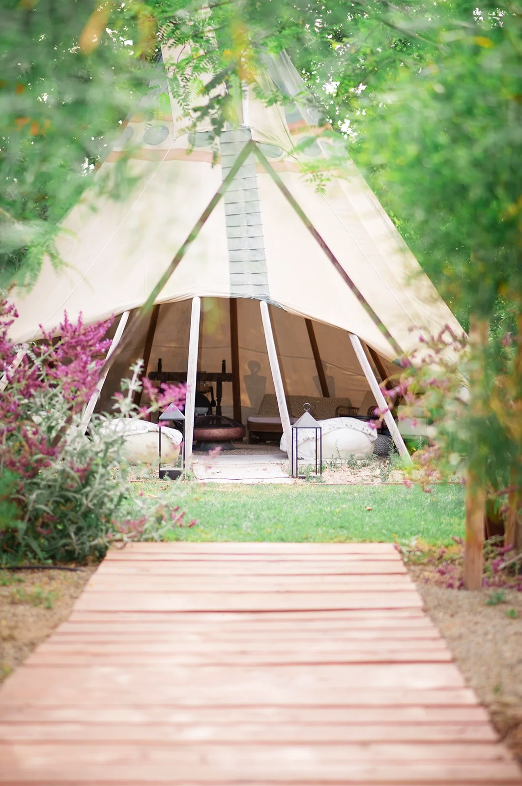 A teepee-style tent with wooden poles, surrounded by greenery and pink flowers, with a wooden walkway leading to it.