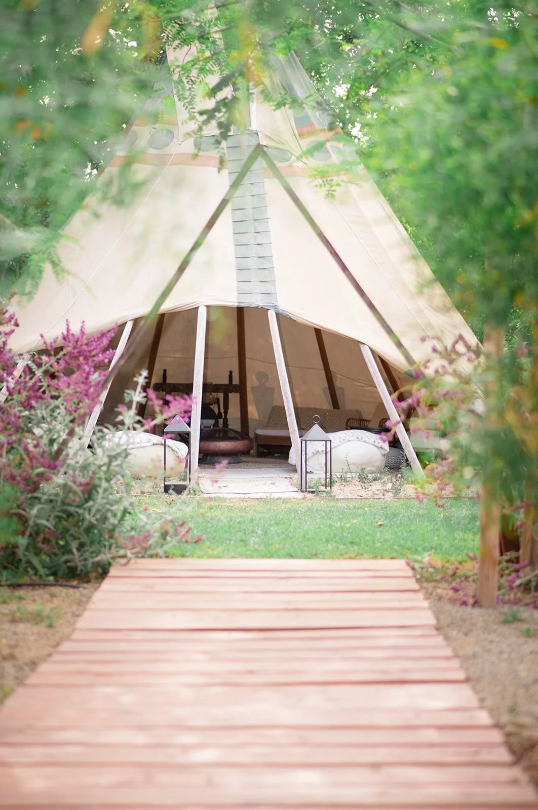 A teepee-style canvas tent set up in a lush garden, viewed through a wooden pathway and surrounded by pink flowers and greenery.