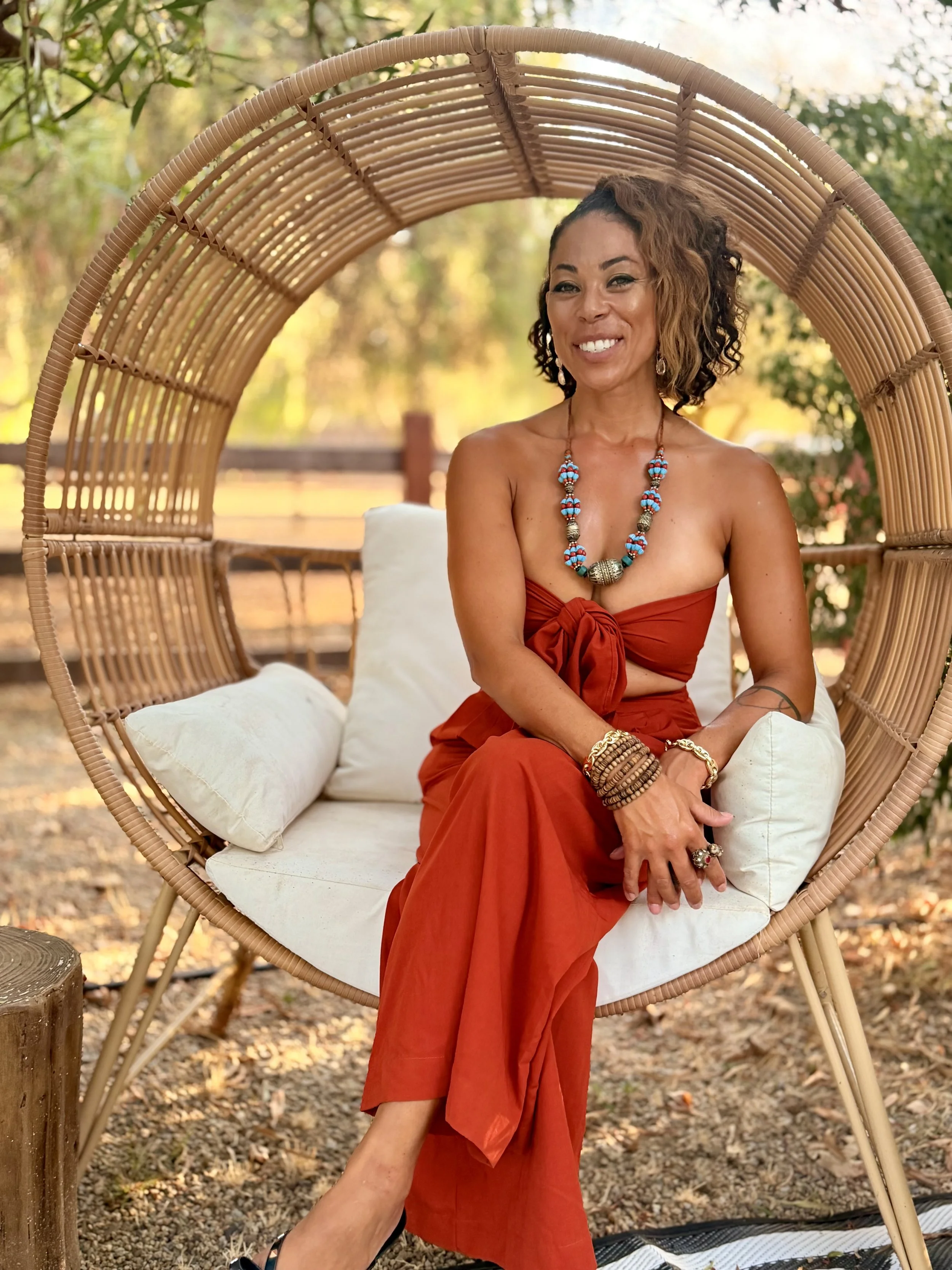 A woman with dark curly hair and a tan complexion sitting on a white cushion inside a circular wicker outdoor chair, smiling, wearing a strapless reddish-orange dress, turquoise and silver bead necklace, and multiple bracelets, outdoors with a blurred background of trees and foliage.