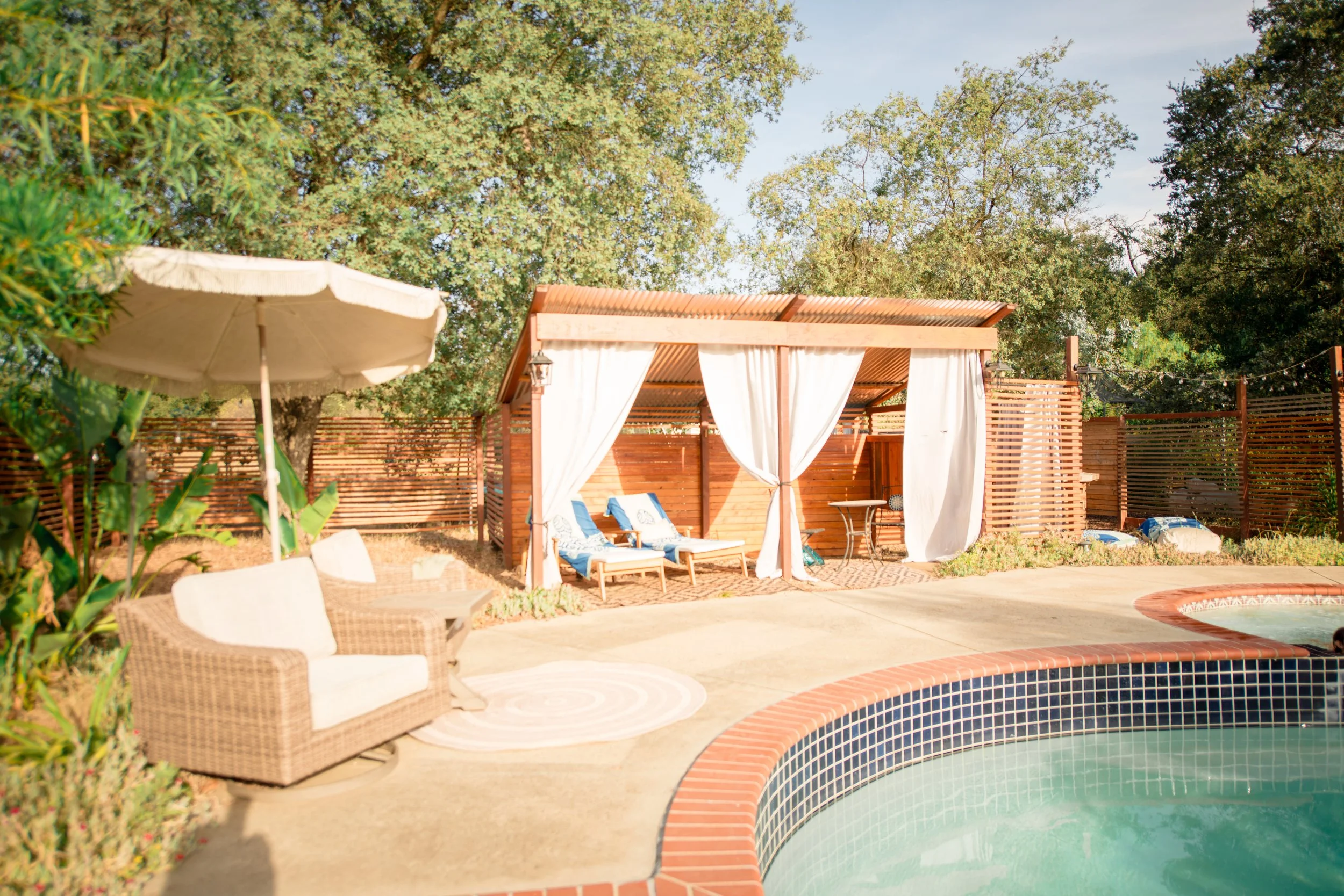 An outdoor backyard scene featuring a swimming pool, a shaded cabana with white curtains, lounge chairs with blue and white cushions, a large beige patio umbrella, and surrounding green trees.