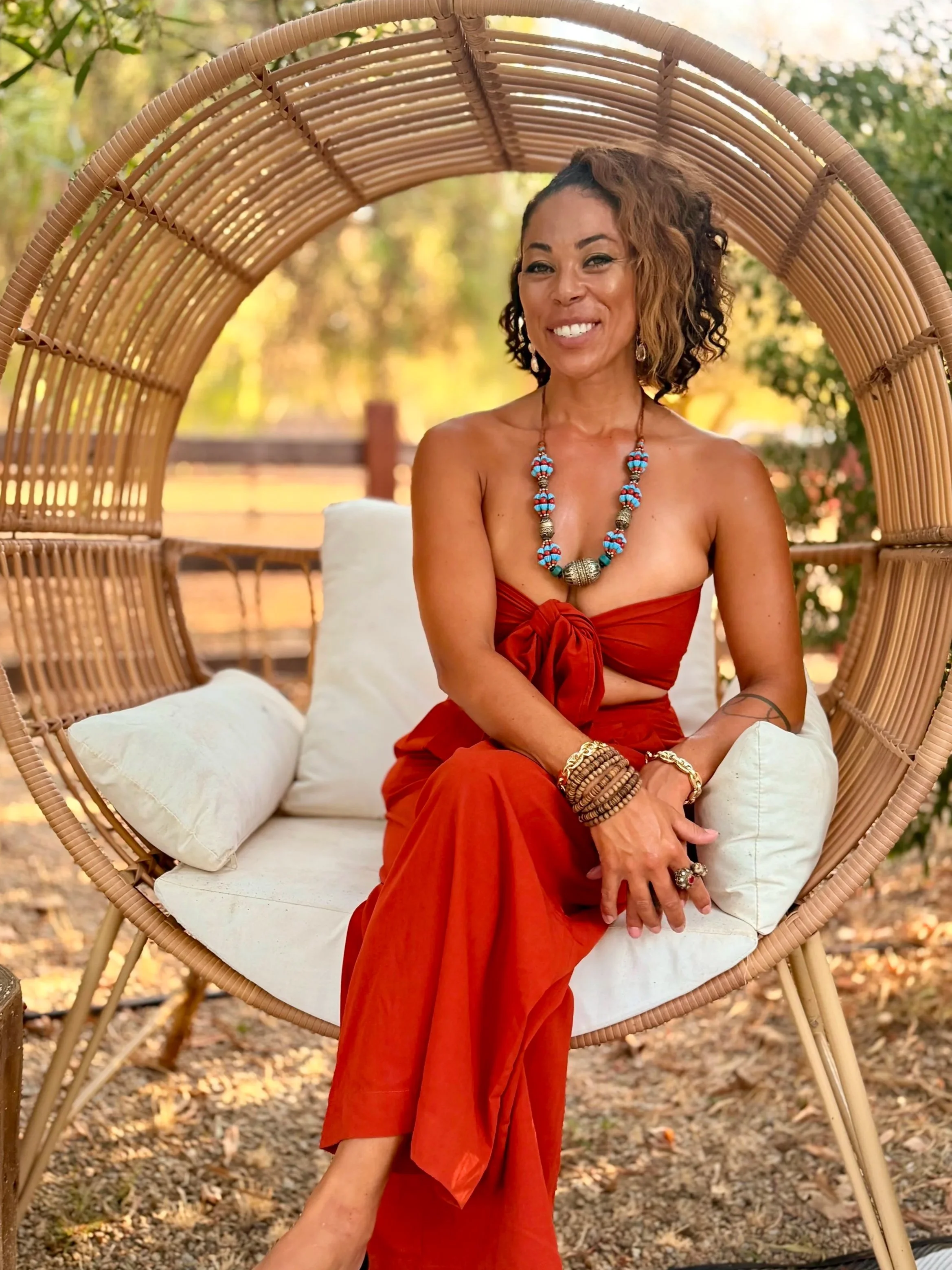 A woman with curly hair and jewelry, wearing a strapless red dress, sitting on a white cushion in a wicker hanging chair outdoors surrounded by trees with autumn leaves.