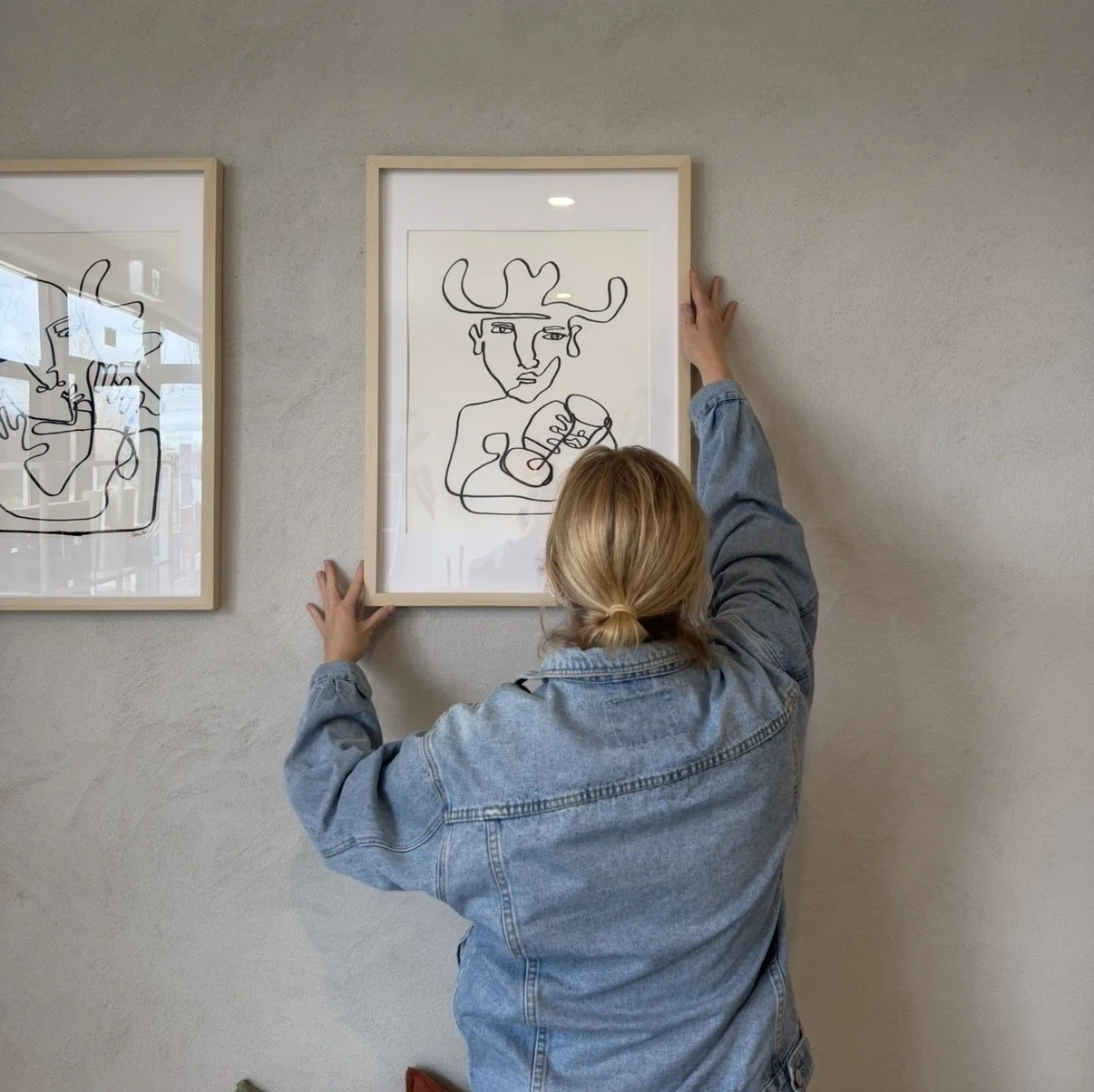 Person hanging a framed line art drawing of a cowboy with a hat, holding a wine glass on a beige wall.