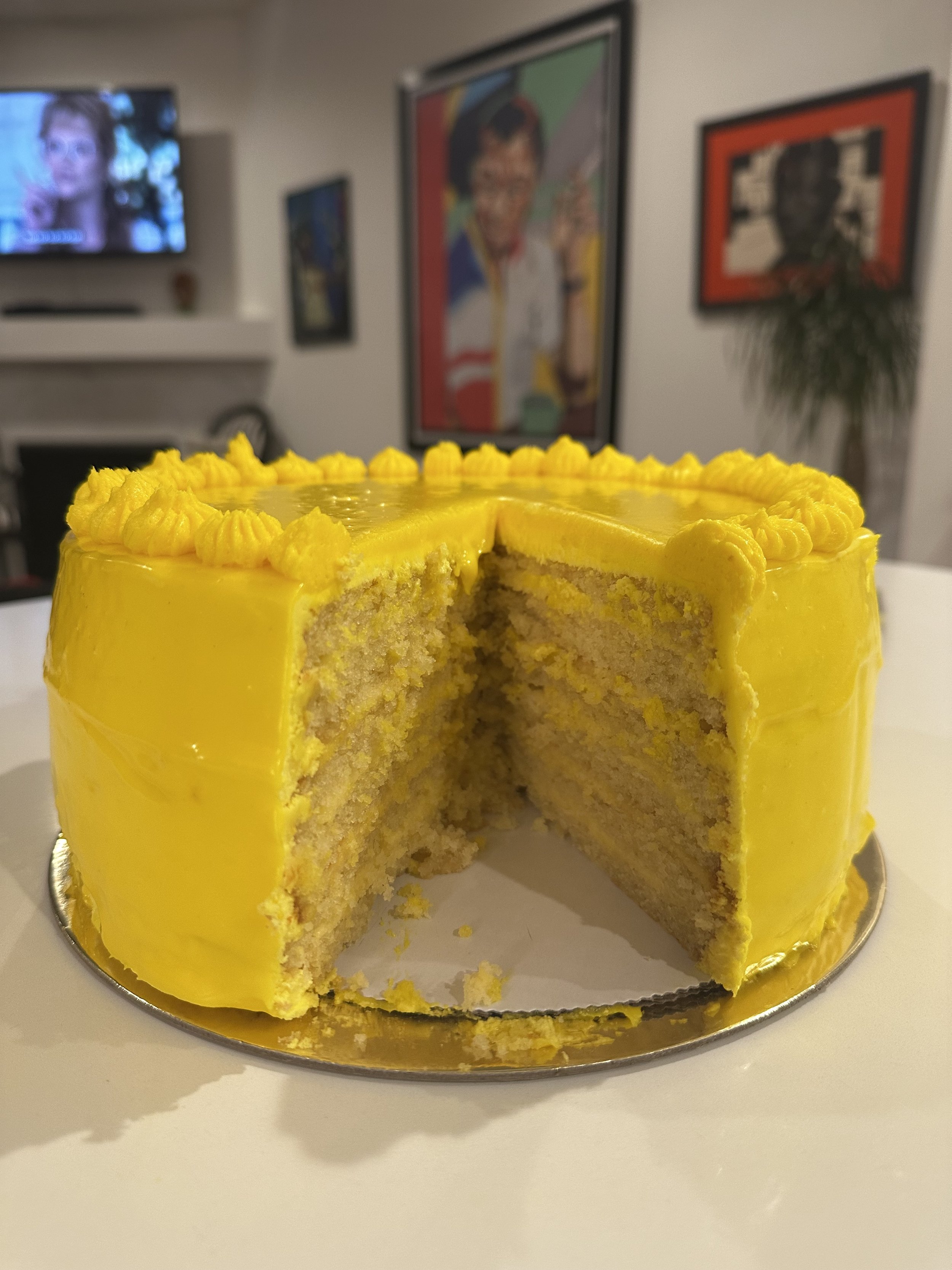 A yellow frosted cake with a slice removed, showing the inside, placed on a silver cake board on a white surface.