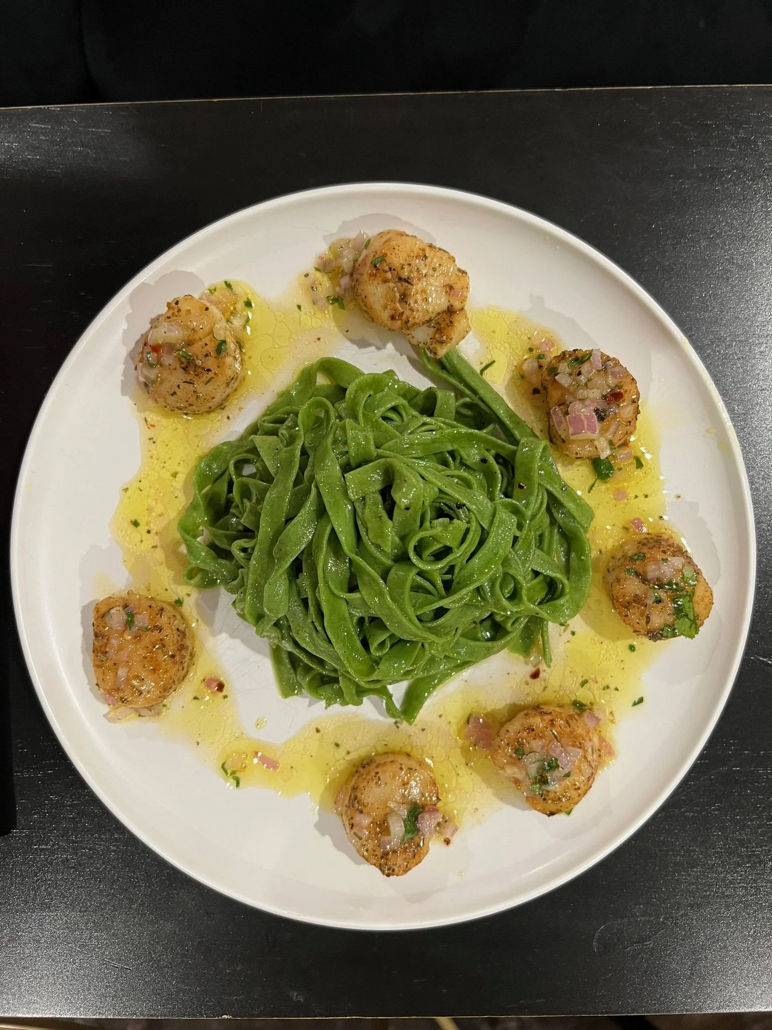 Plate with cooked green pasta noodles and several cooked scallops topped with chopped herbs and small bits of onion, drizzled with olive oil.