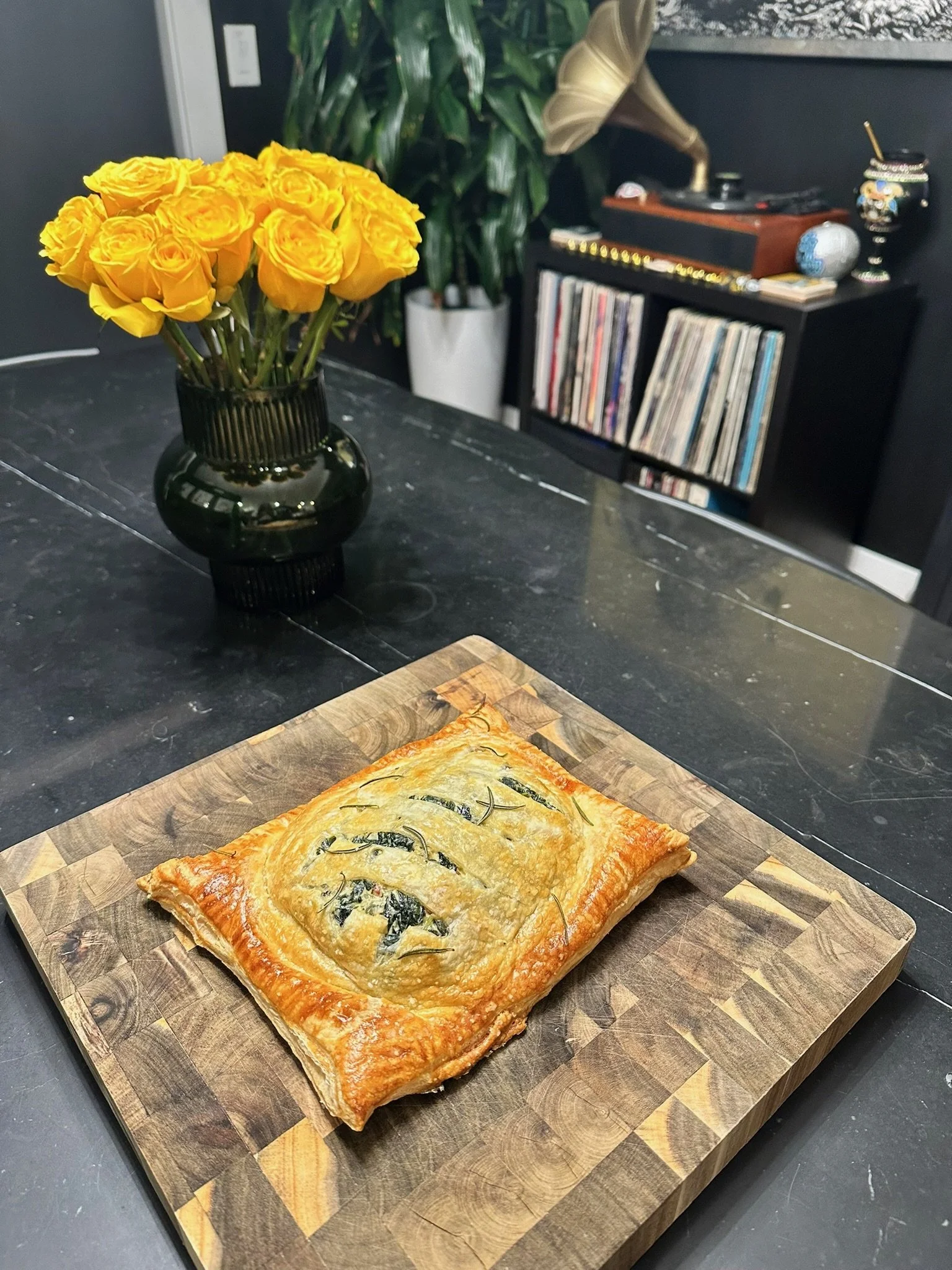 A vegetable tart with a golden-brown crust, herbs, and greens on a wooden cutting board on a black countertop.